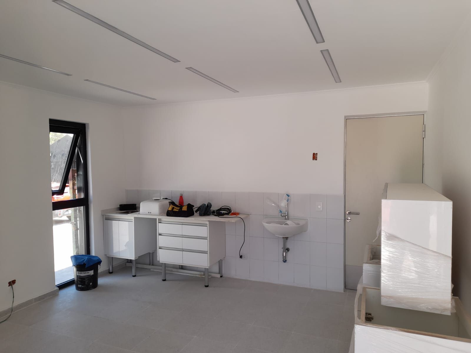 Habitación blanca vacía con equipo médico, lavabo, puerta y ventana.