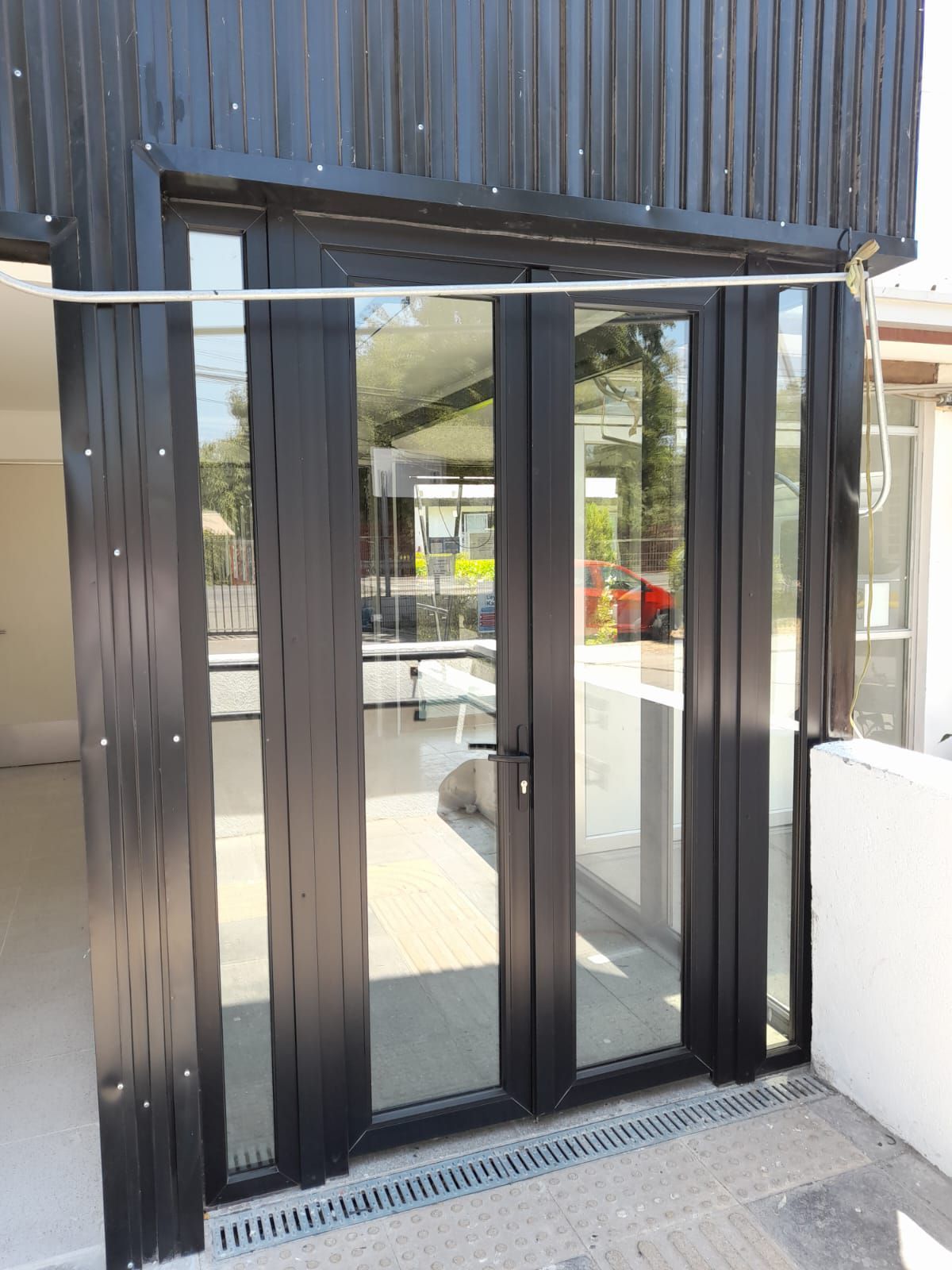 Las puertas de vidrio con marco negro se abren a un interior luminoso. El exterior cuenta con revestimiento negro y canal de drenaje.