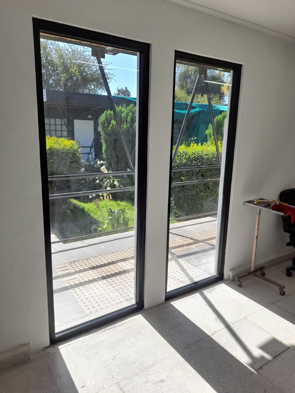 Dos ventanas altas con marco negro; la luz del sol entra a raudales en una habitación de paredes blancas, reflejando un patio verde y un edificio exterior.