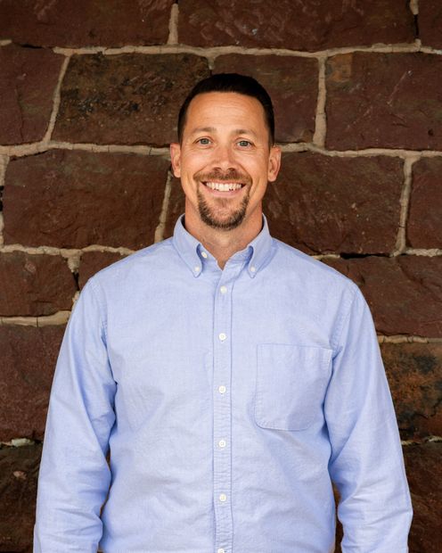Man with short dark hair and goatee smiles in front of a brown stone wall, wearing a purple plaid button-down shirt.