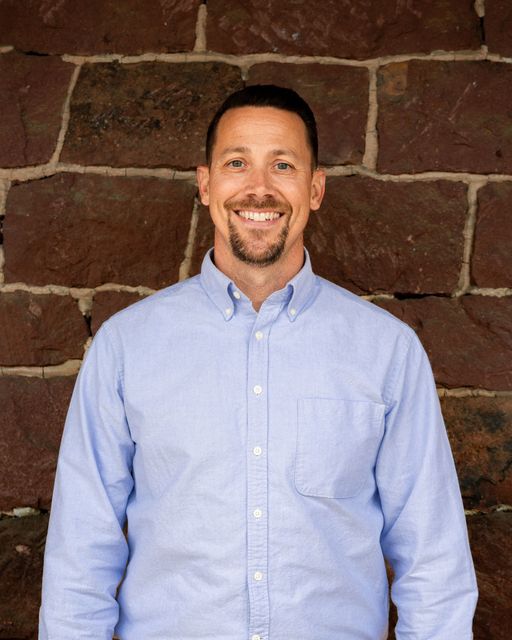 Man with short dark hair and goatee smiles in front of a brown stone wall, wearing a purple plaid button-down shirt.