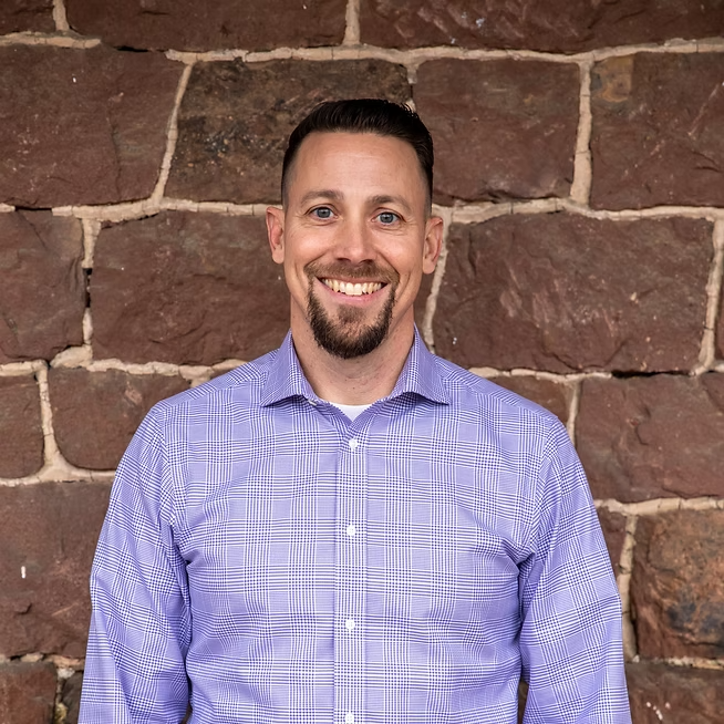 Man with short dark hair and goatee smiles in front of a brown stone wall, wearing a purple plaid button-down shirt.