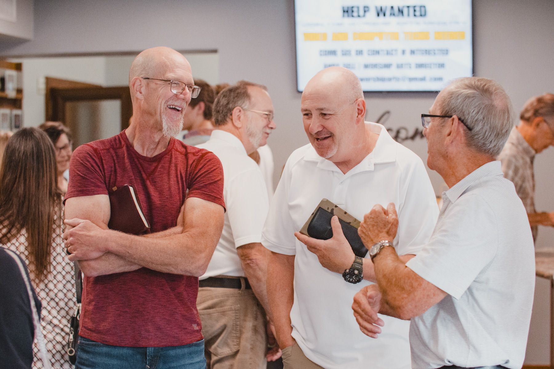Three older men conversing in a room; two wear white shirts, one a maroon shirt. A 