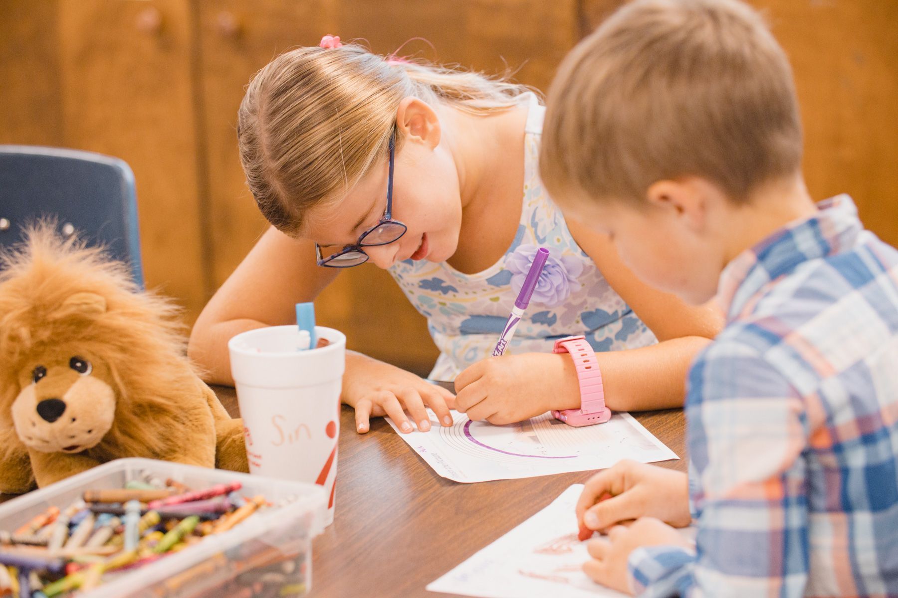 Girl with glasses and boy drawing at a table, lion stuffed animal nearby.