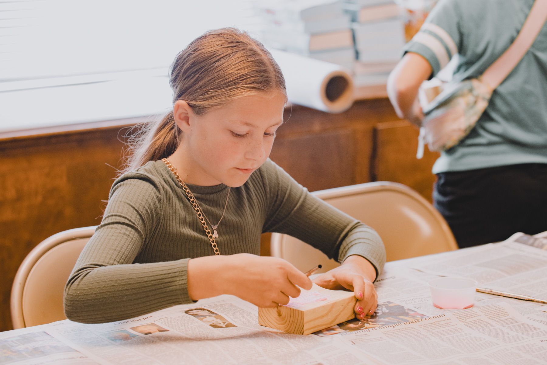 Girl in green shirt working on a craft project at a table with a second person standing nearby.