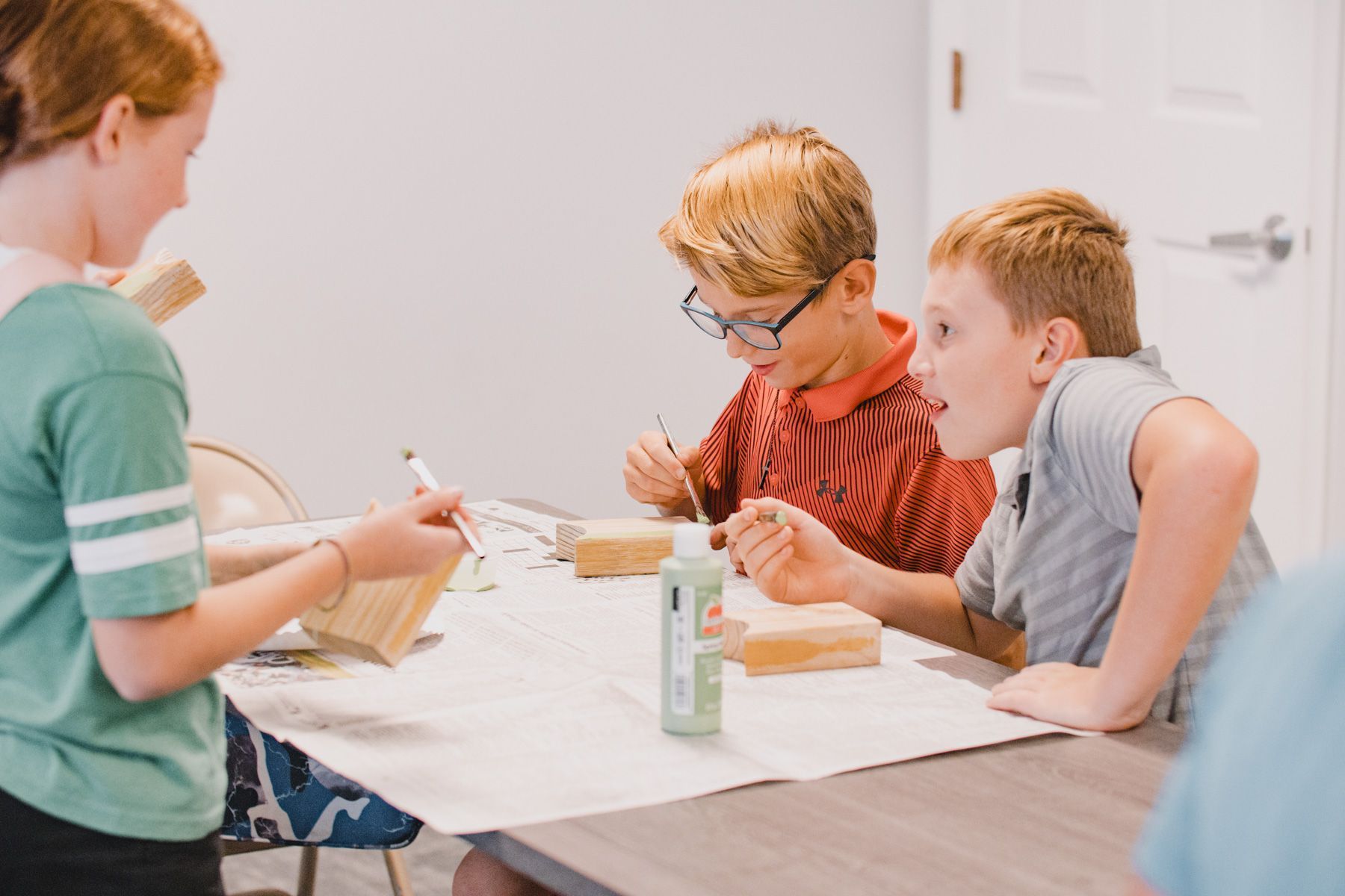 Three children painting wooden objects at a table; light interior.