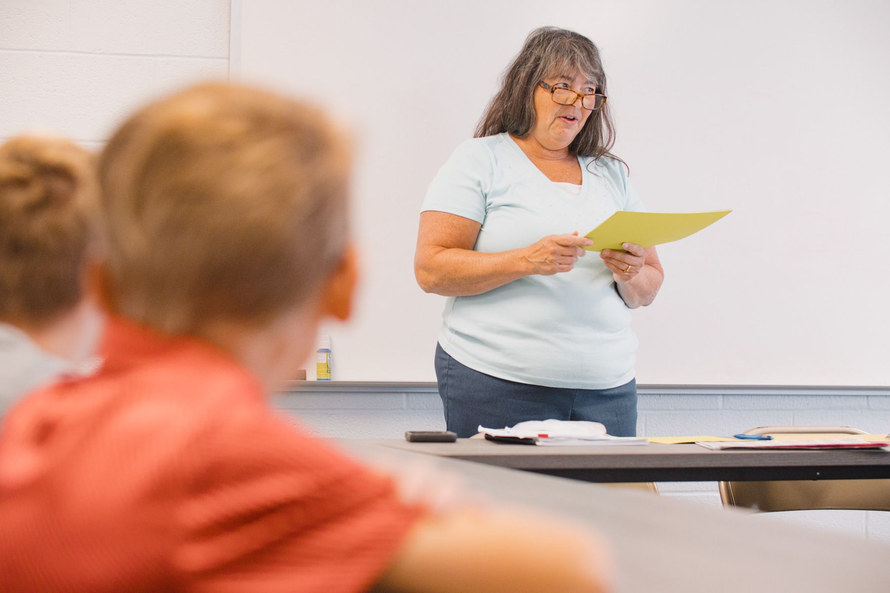 Teacher stands at the front of a classroom reading from a yellow paper, facing students. Whiteboard in background.