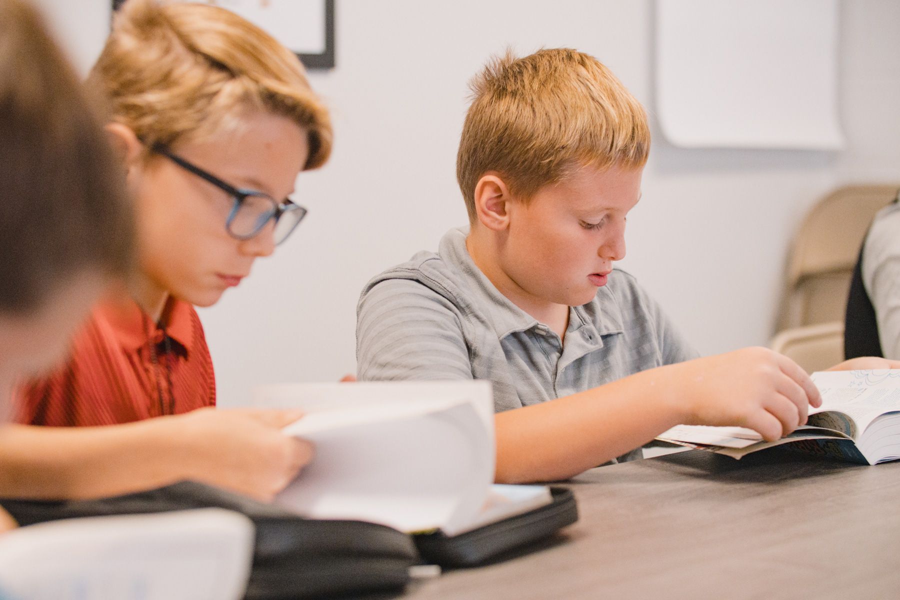 Two boys reading books at a table indoors. One wears glasses, the other a grey shirt.