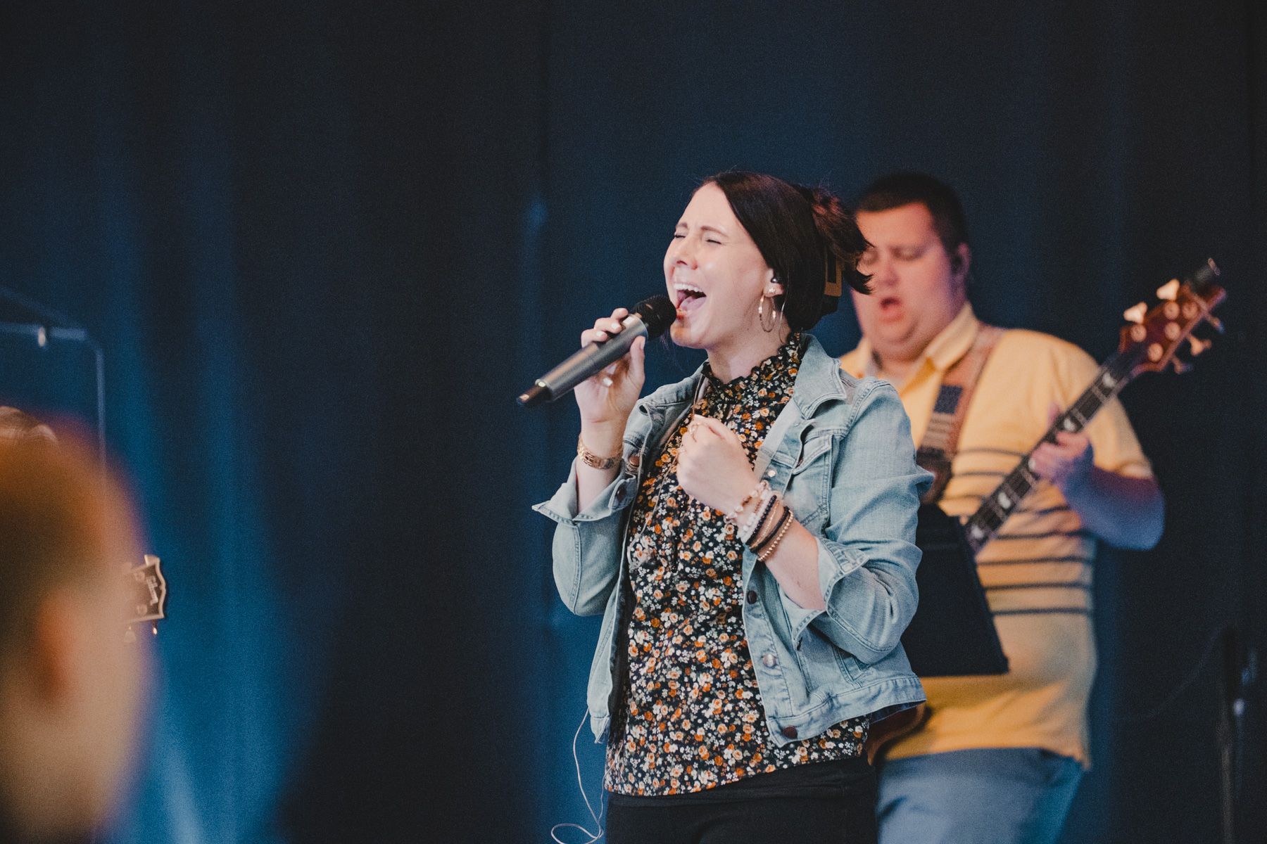 Woman singing into a microphone on stage; man plays guitar in the background.