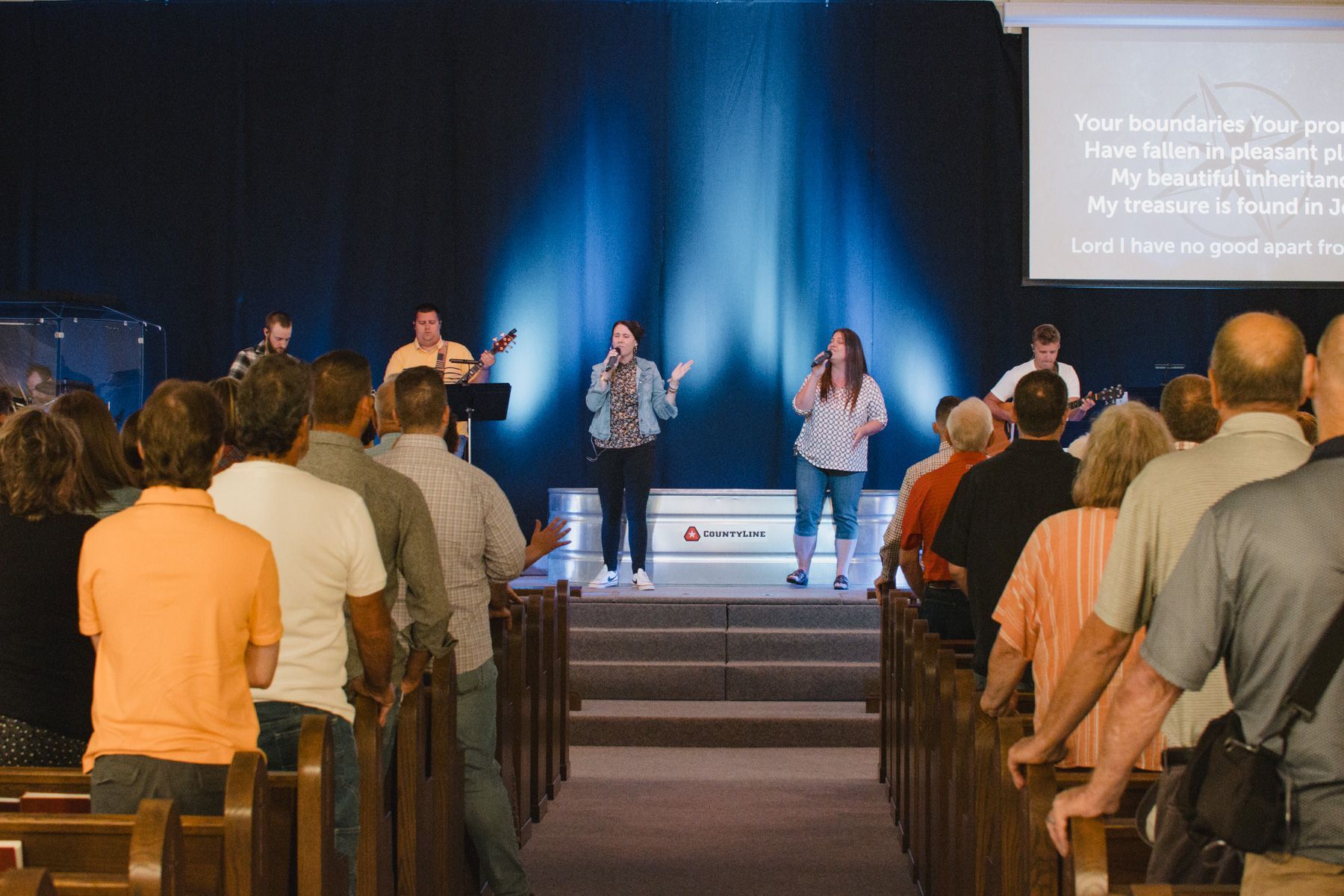 People singing and listening in a church. Stage lit with blue light, two women singing with band, rows of pews.