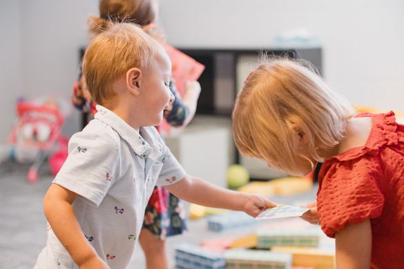 A young boy smiles and hands a card to a girl in a playroom.