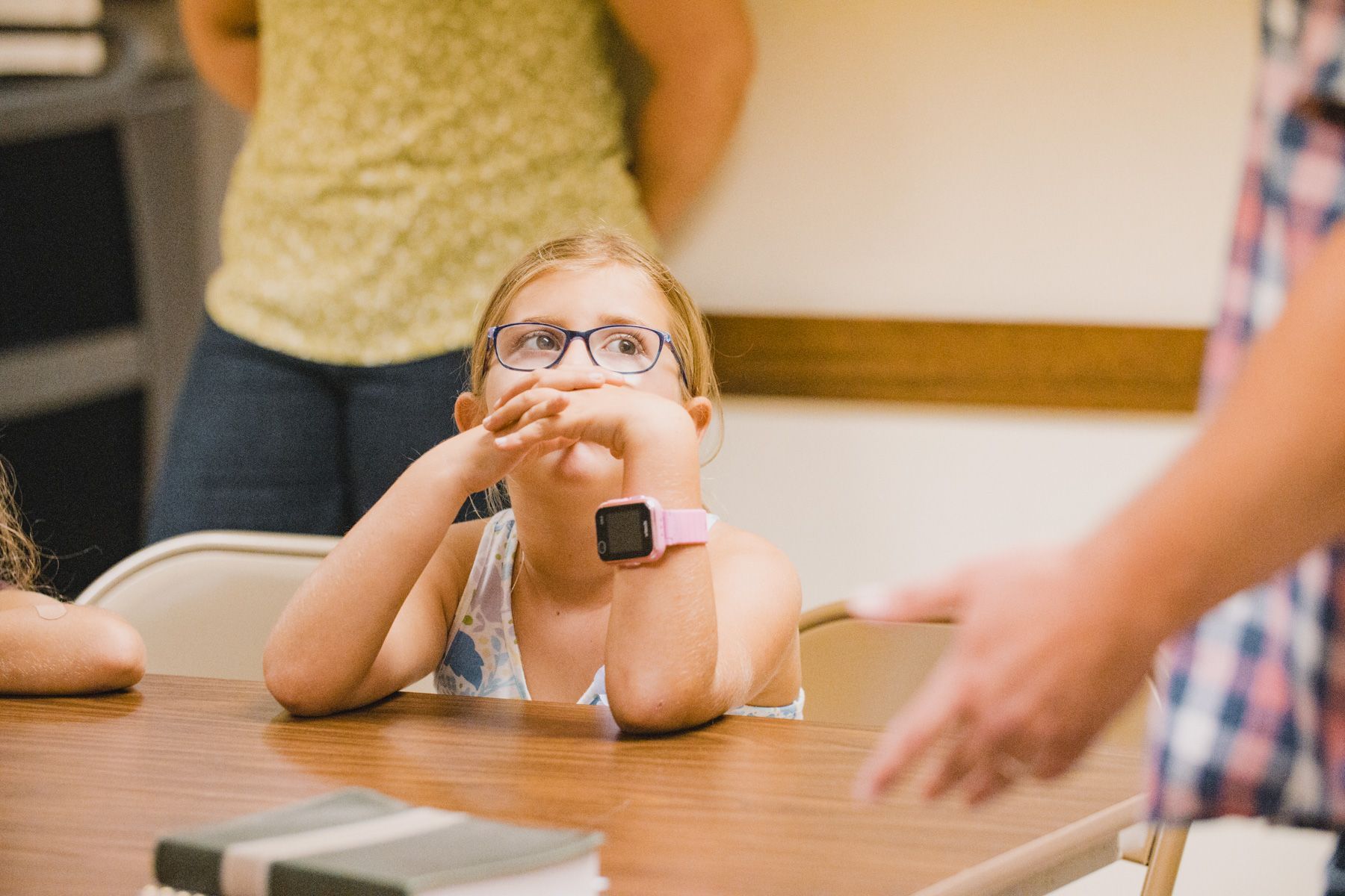 Girl with glasses leans on table, looking up, hands near her face, in a room with people.