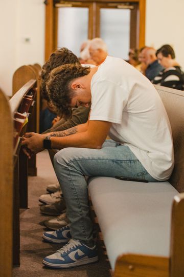 Young man in light jeans and a white t-shirt prays with head bowed in a church pew.