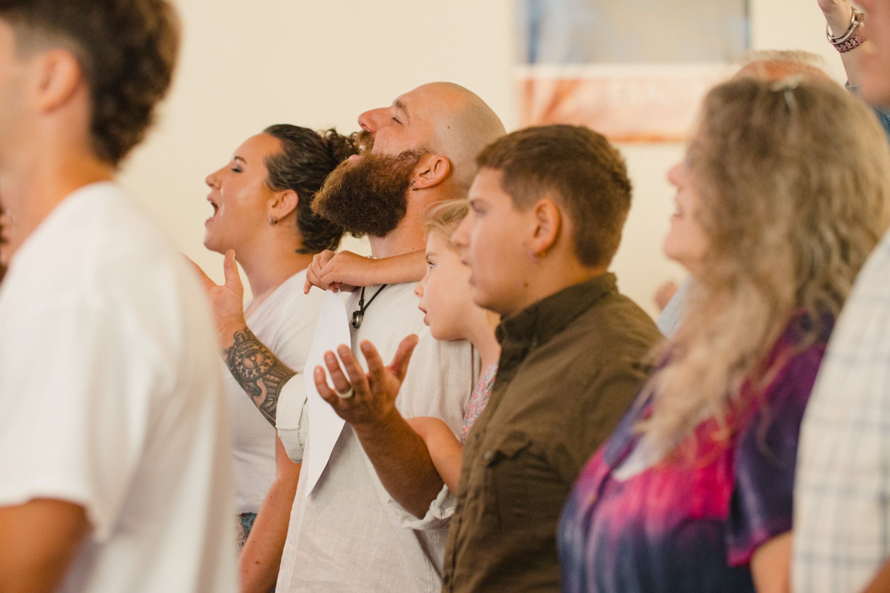 People worshiping with arms raised, some looking upwards, inside a building with natural light.