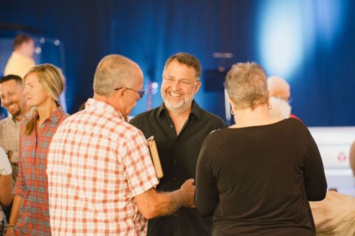 Man shaking hands with another, smiling. Several people in a room, blue background.