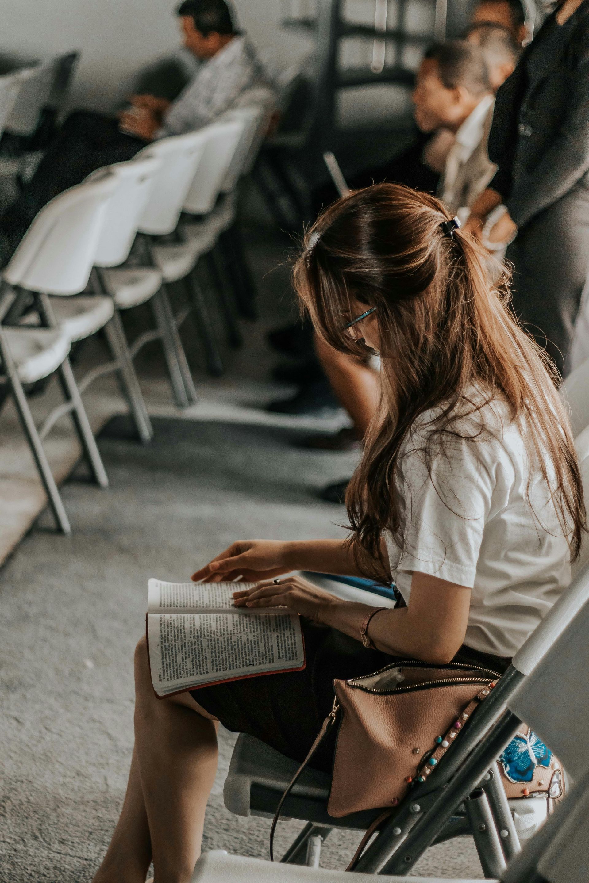 Woman with long hair, reading a book while seated in a room with other people.