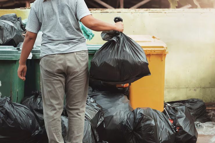Person throwing a black trash bag into a yellow dumpster, surrounded by more bags.