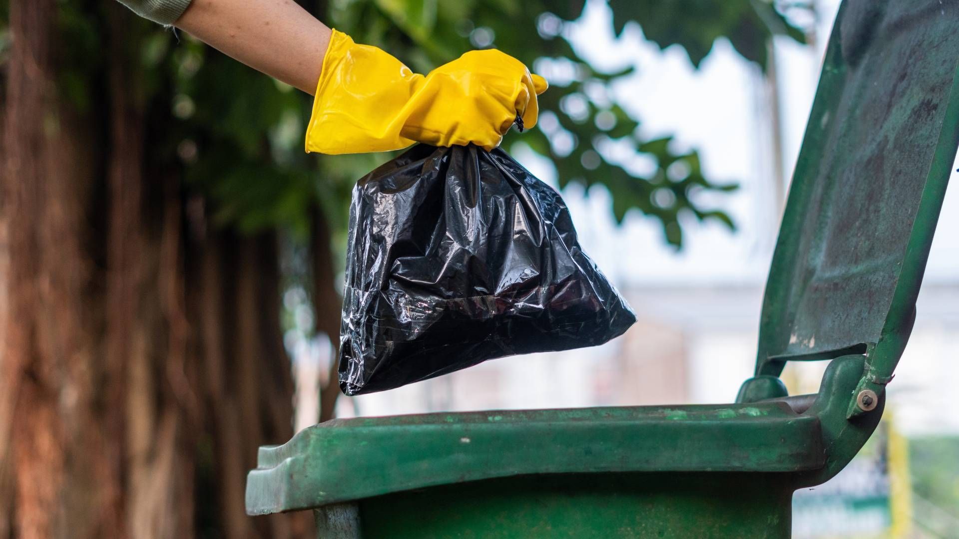 Hand in yellow glove throwing a black trash bag into a green garbage can.