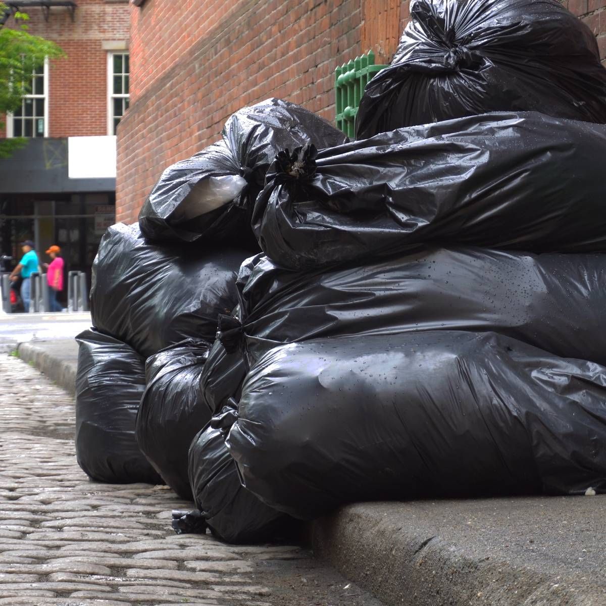 Pile of black trash bags on a city sidewalk, near a brick building.