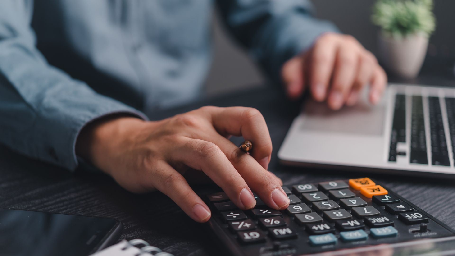 A person is using a calculator in front of a laptop computer.