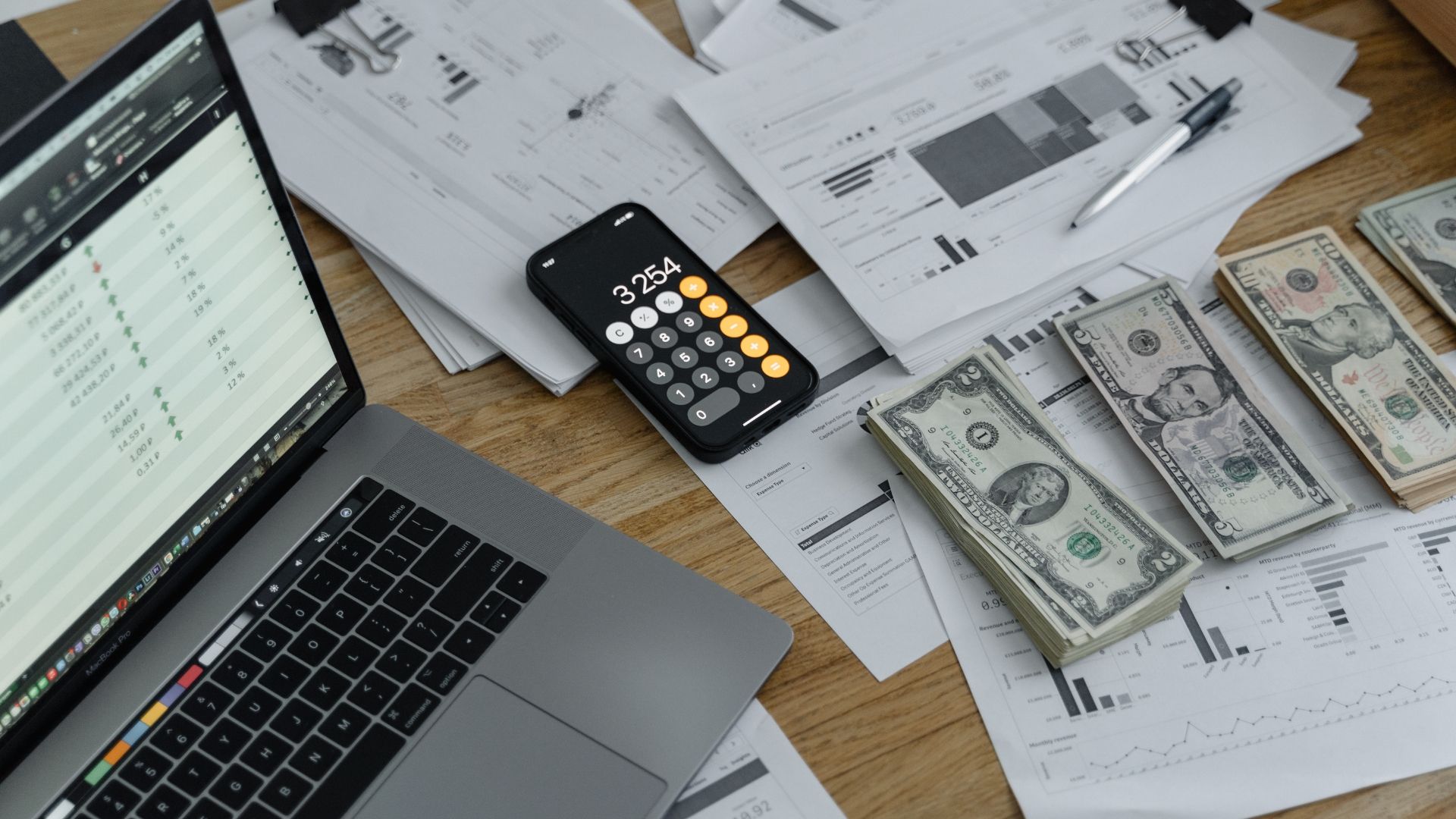 A laptop , cell phone , calculator , and stacks of money are on a wooden table.