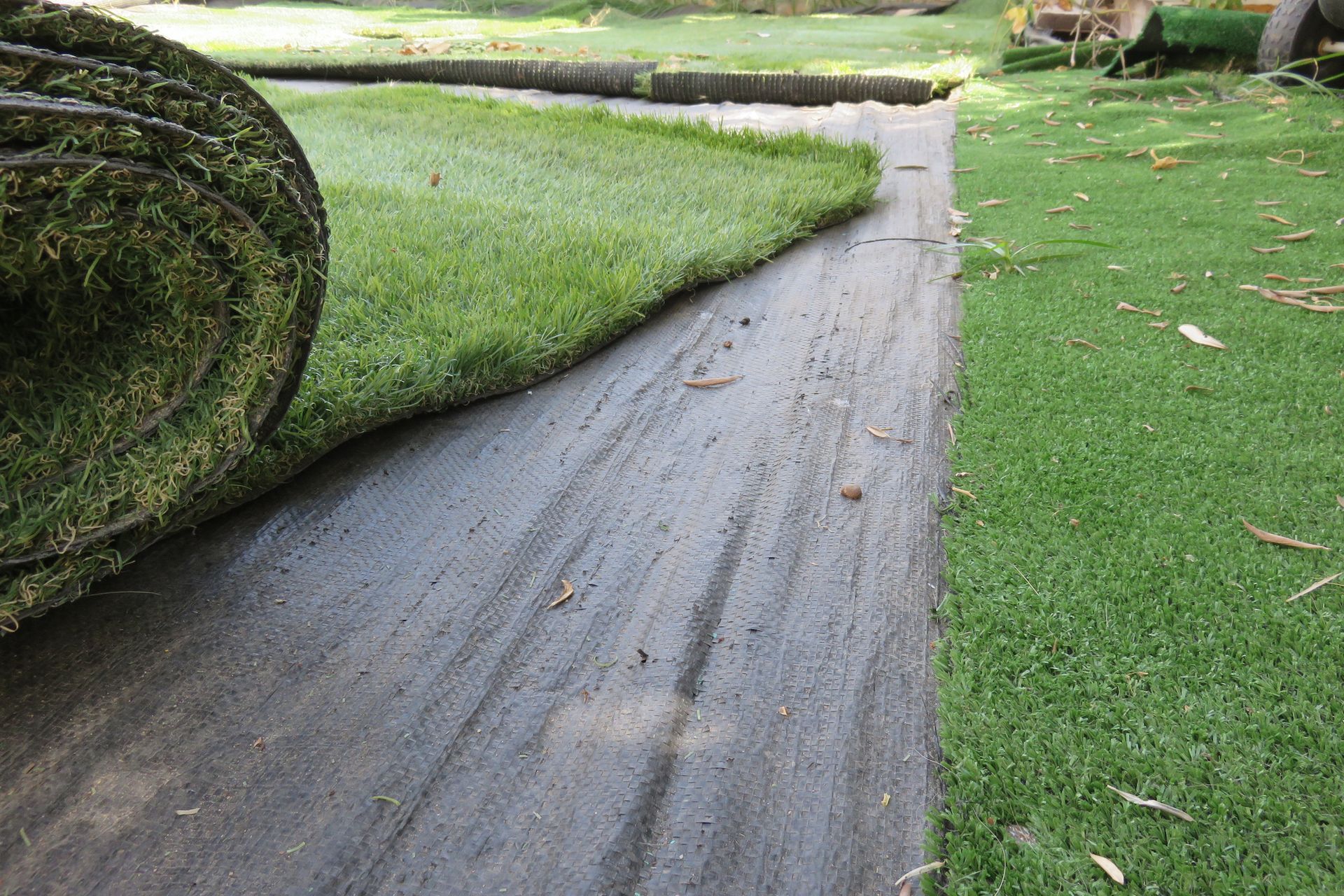 A roll of artificial turf being unrolled onto a black underlayment next to an already-laid section of green turf in a yard.