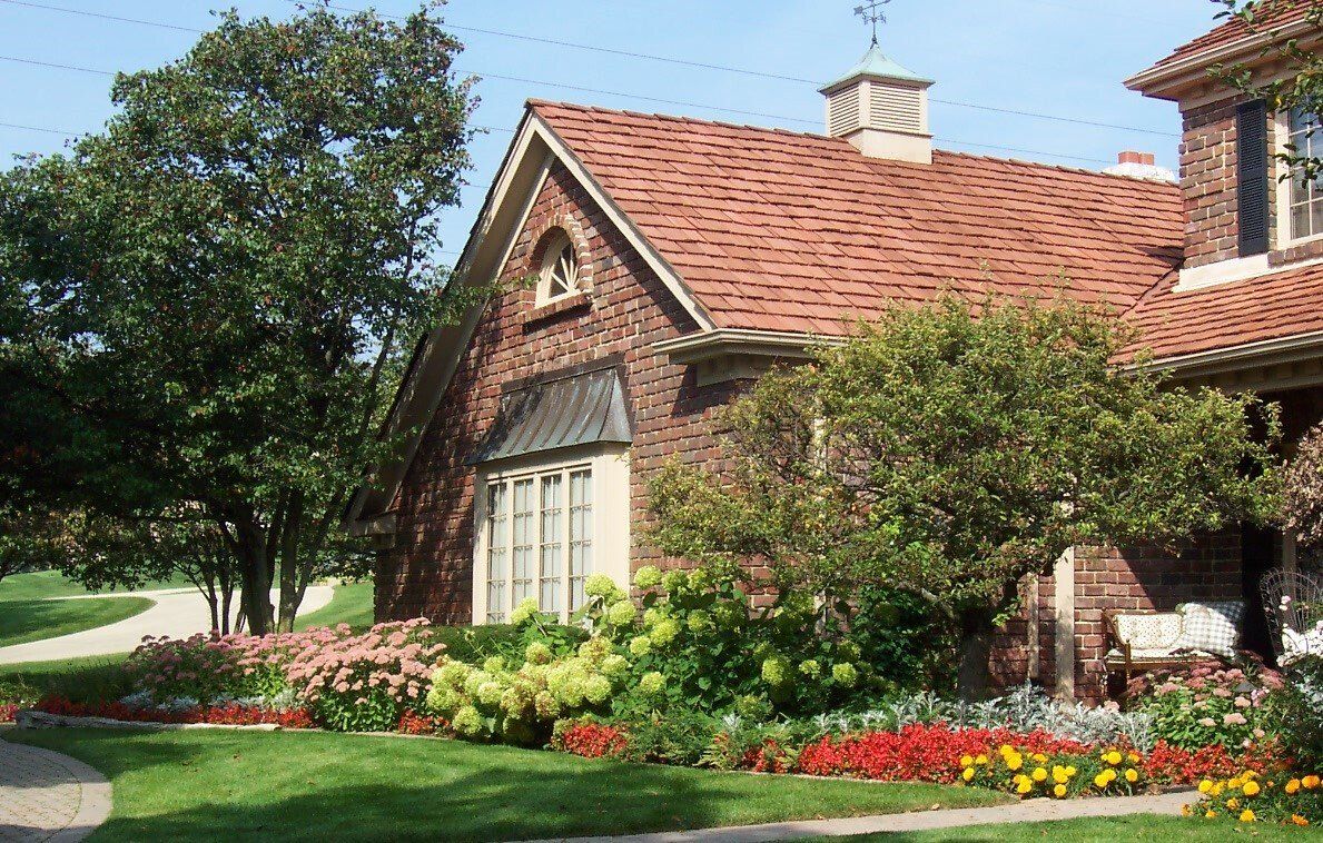 A brick house with a red roof is surrounded by flowers and trees