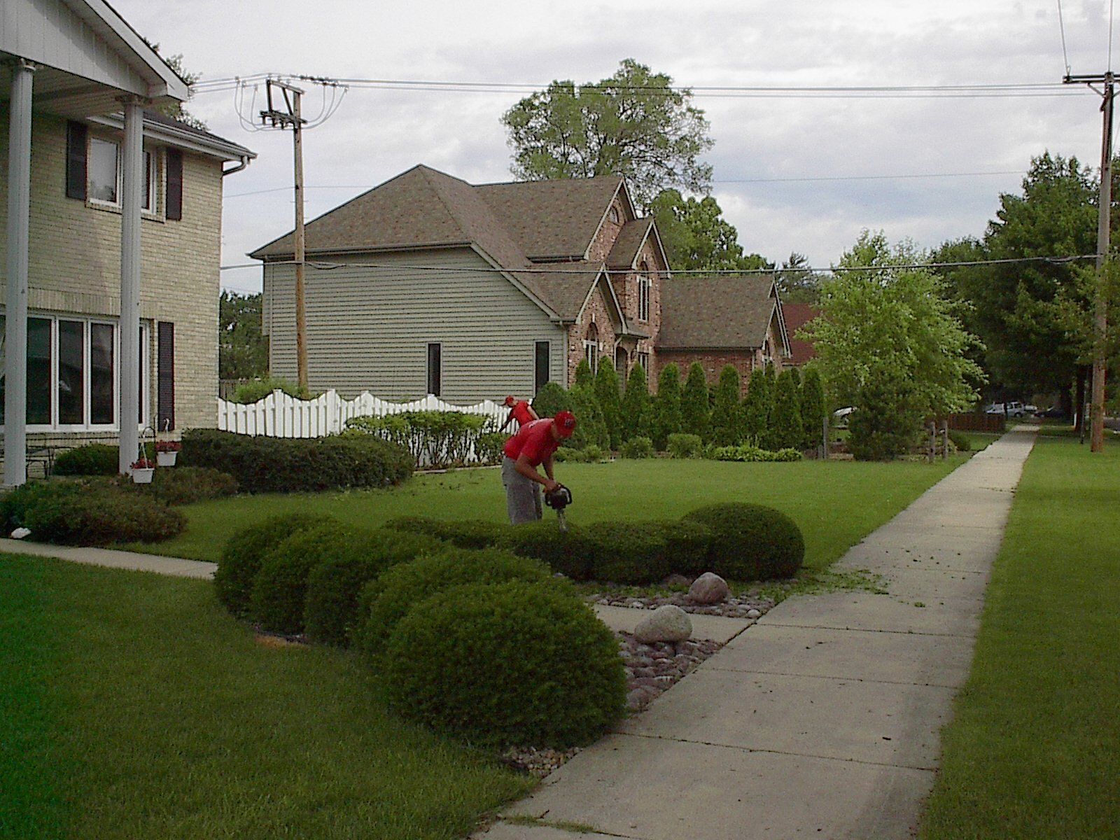 A man in a red shirt is cutting bushes in front of a house