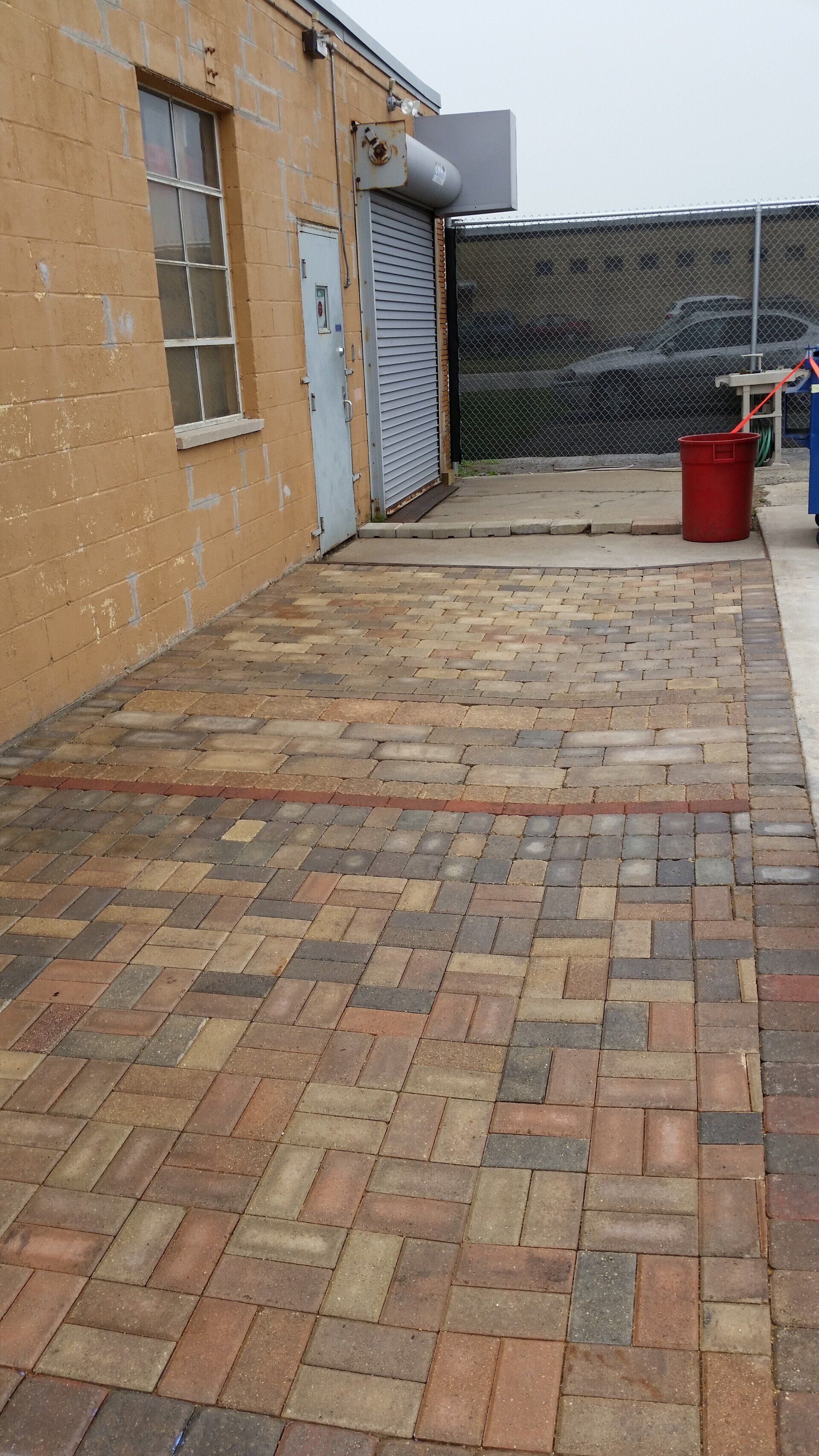 A brick walkway leading to a building with a red trash can in front of it.
