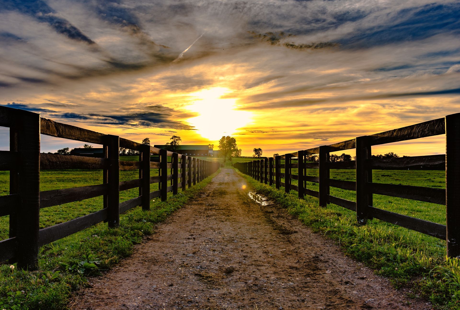 The sun is setting over a dirt road lined with wooden fences.