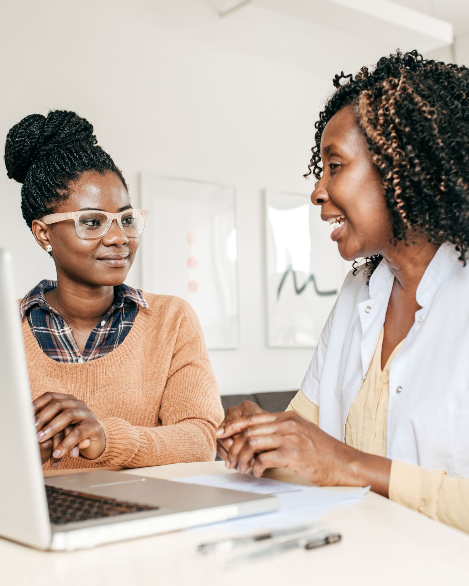 Woman speaking with another woman in a medical setting, both smiling. A laptop and paperwork are on the desk.