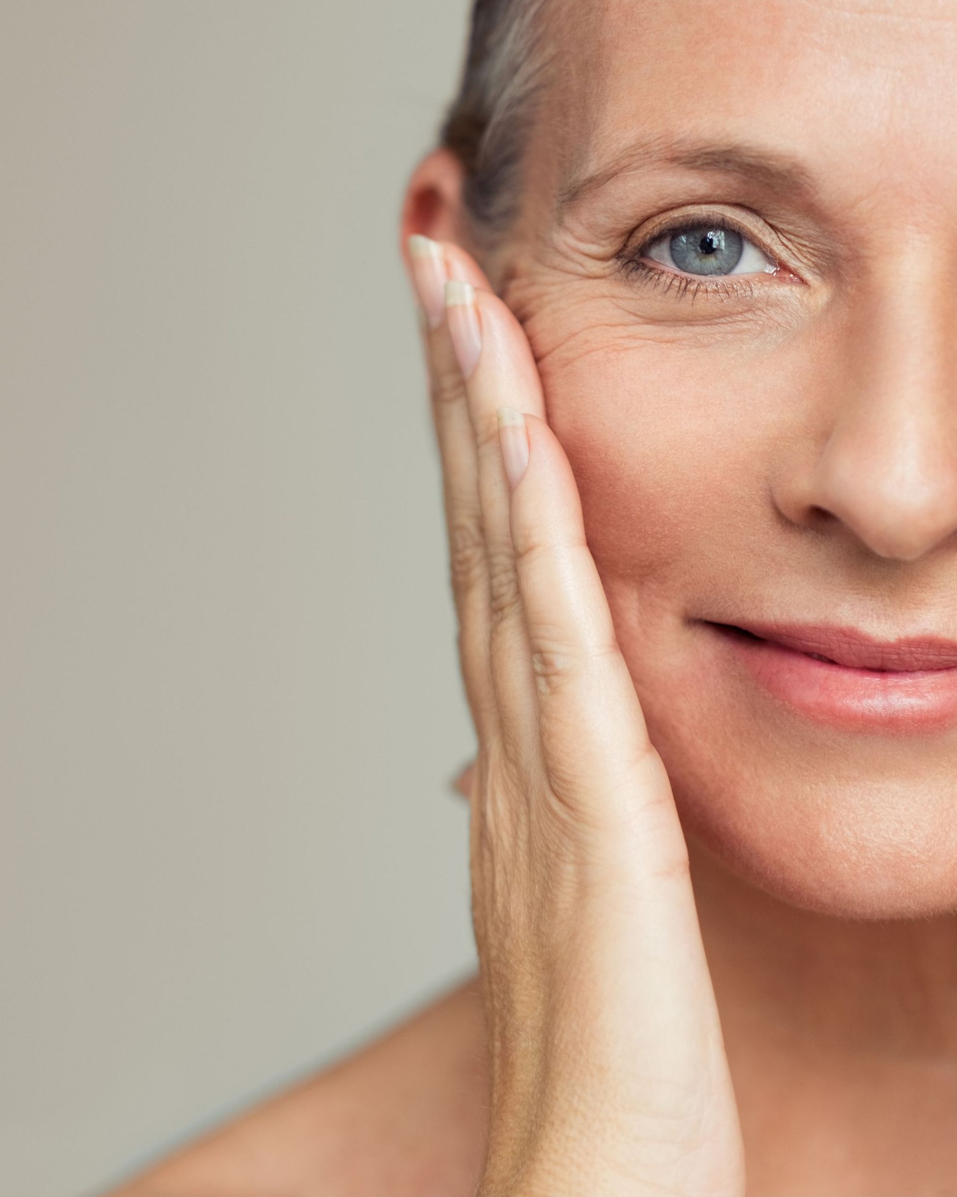 Close-up of a person's face with wrinkles, touching their cheek with their hand, against a neutral background.