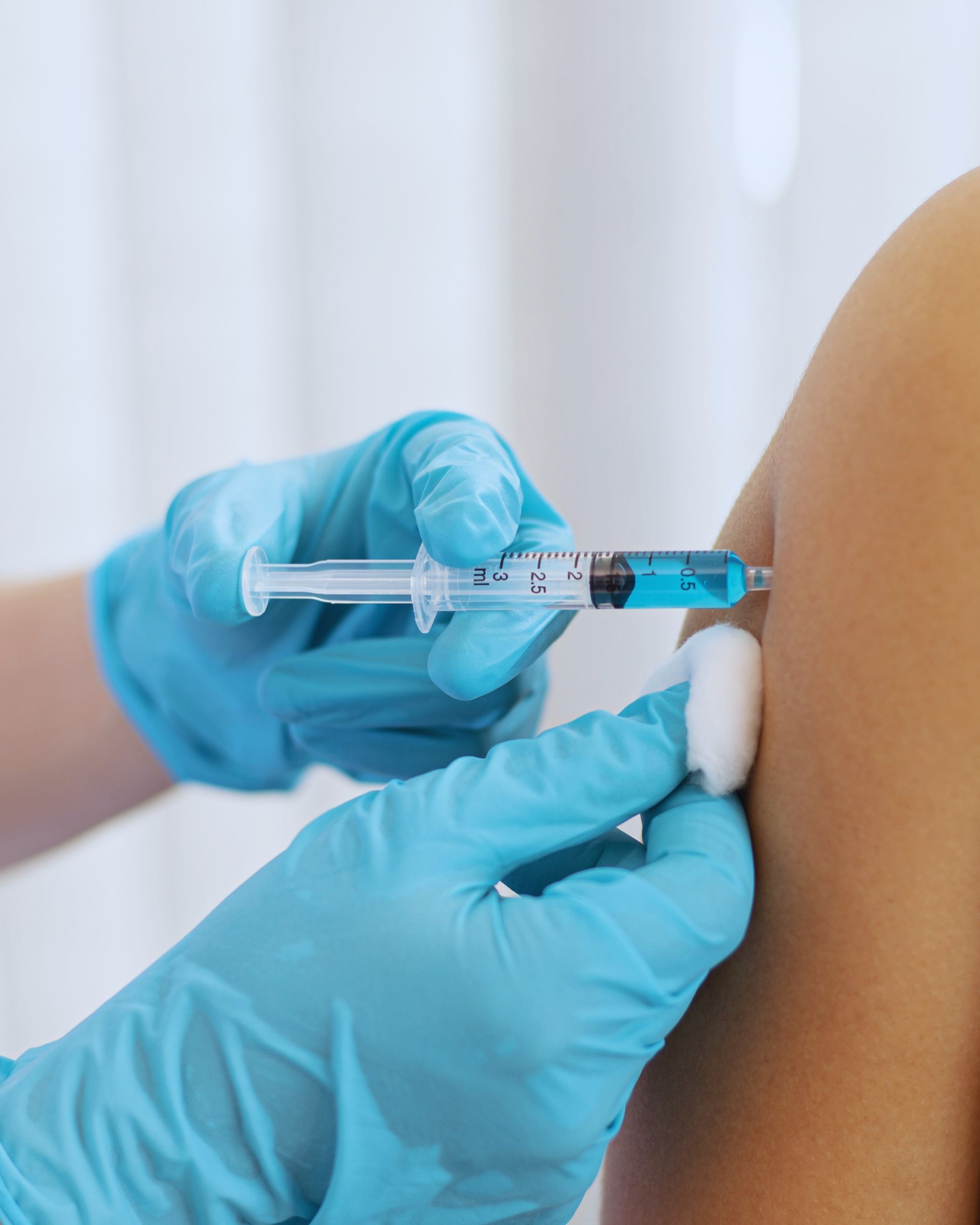Person in blue gloves administering an injection with a syringe into an arm, held with cotton.