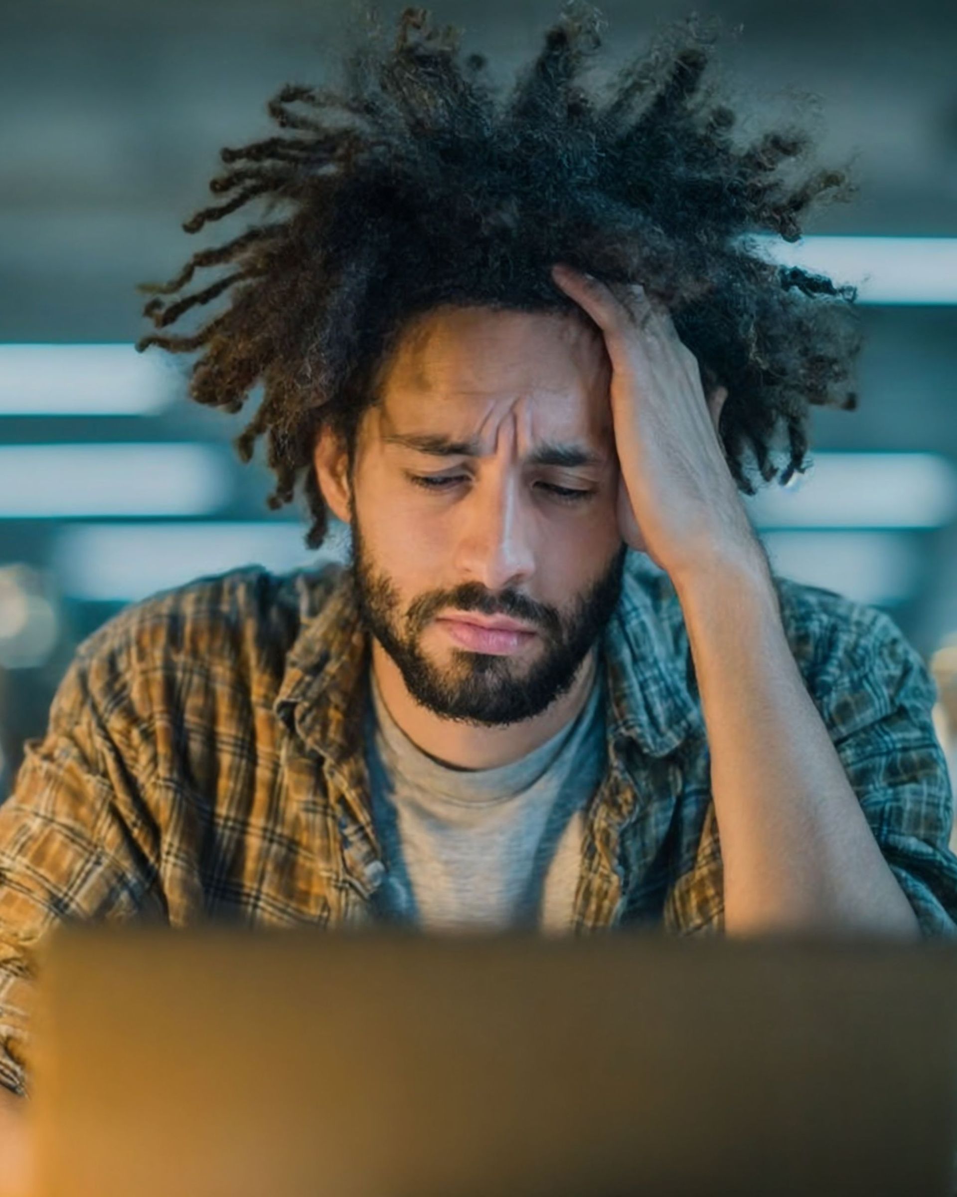 Man with dreadlocks holds his head, looking stressed, at a laptop.