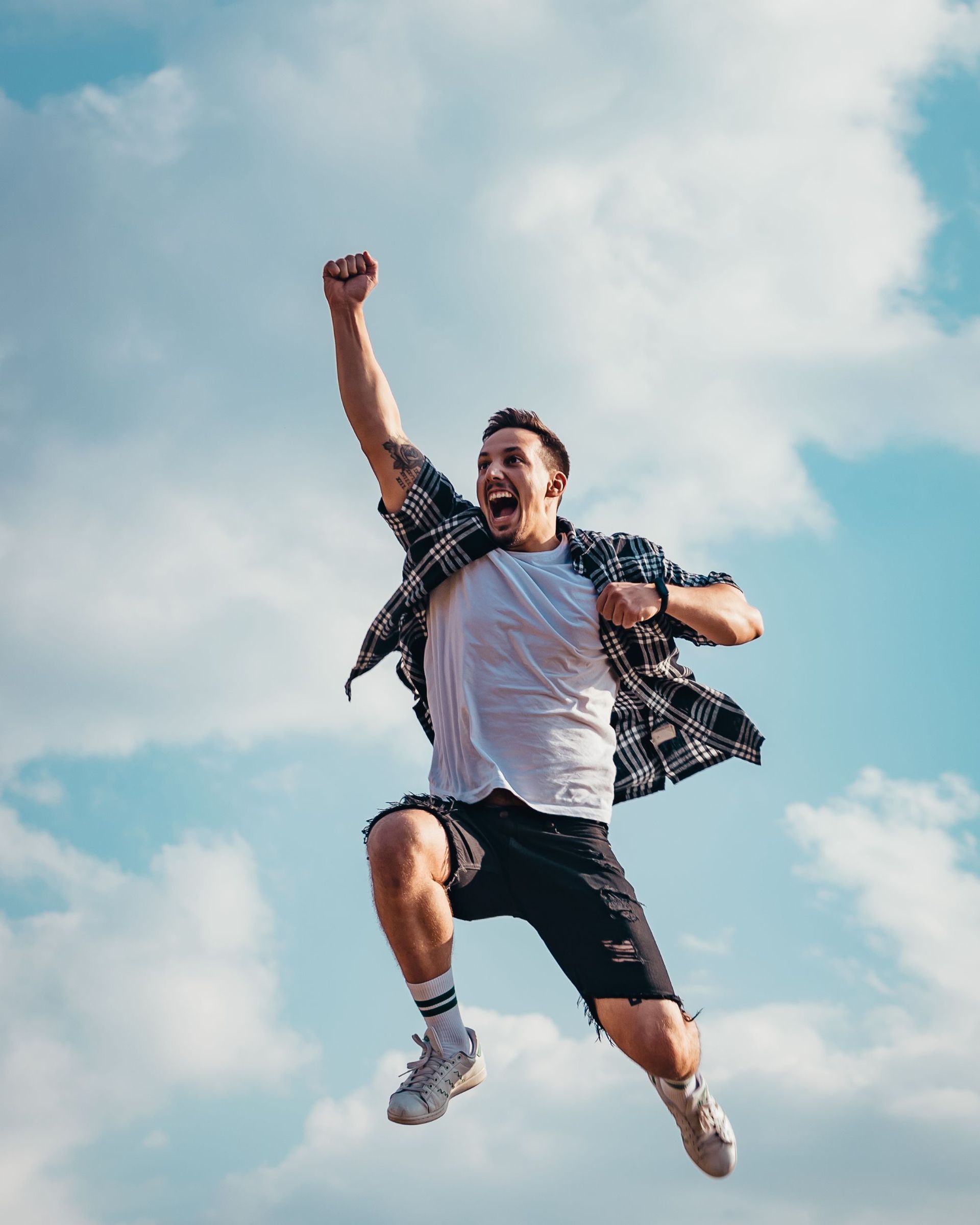 Man jumps with fist raised in the air, open mouth expression, against a blue and cloudy sky.