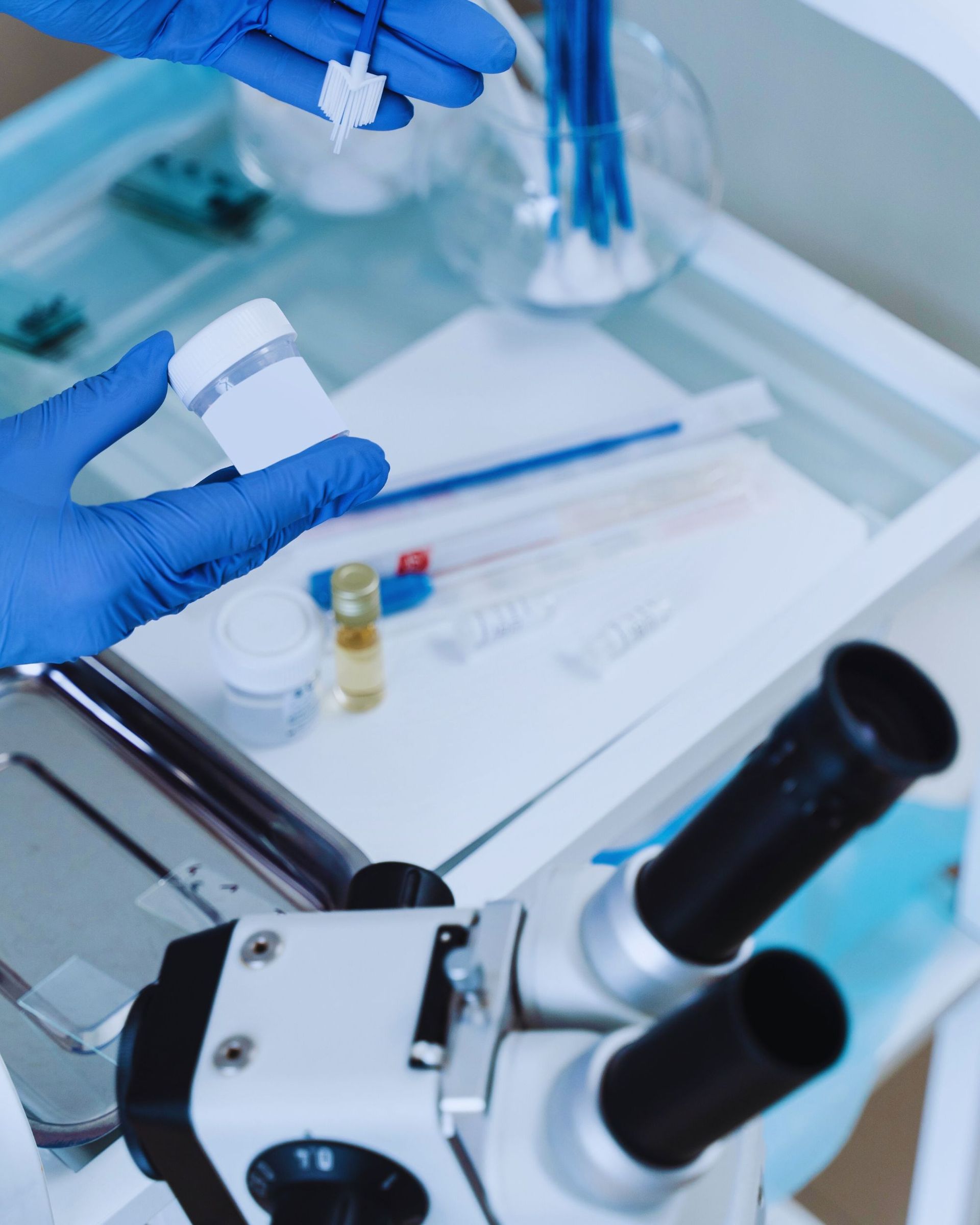 Lab worker with blue gloves holds a container and small sample; microscope and lab supplies visible.