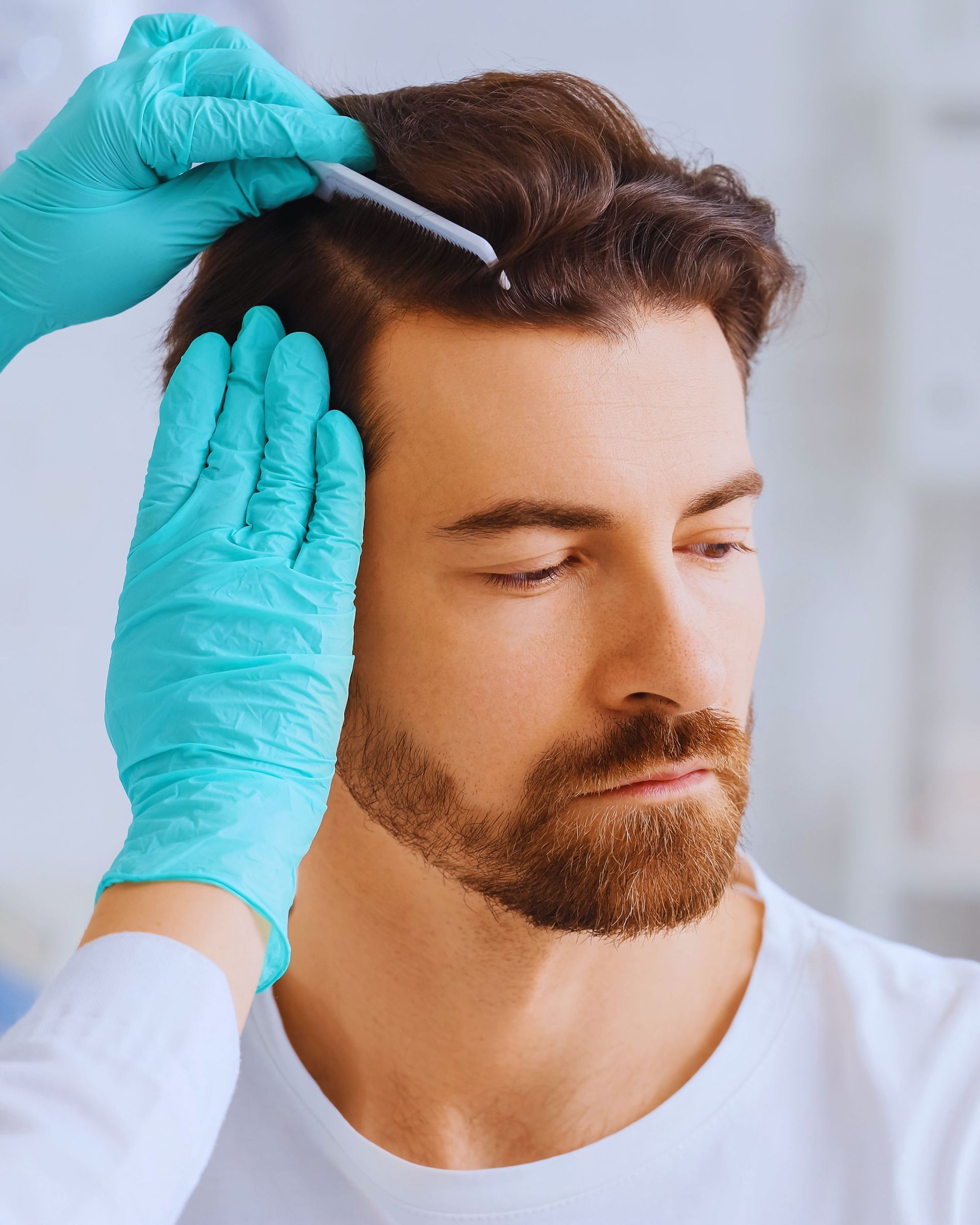 Man receiving a hair treatment; gloved hands part hair with a tool.