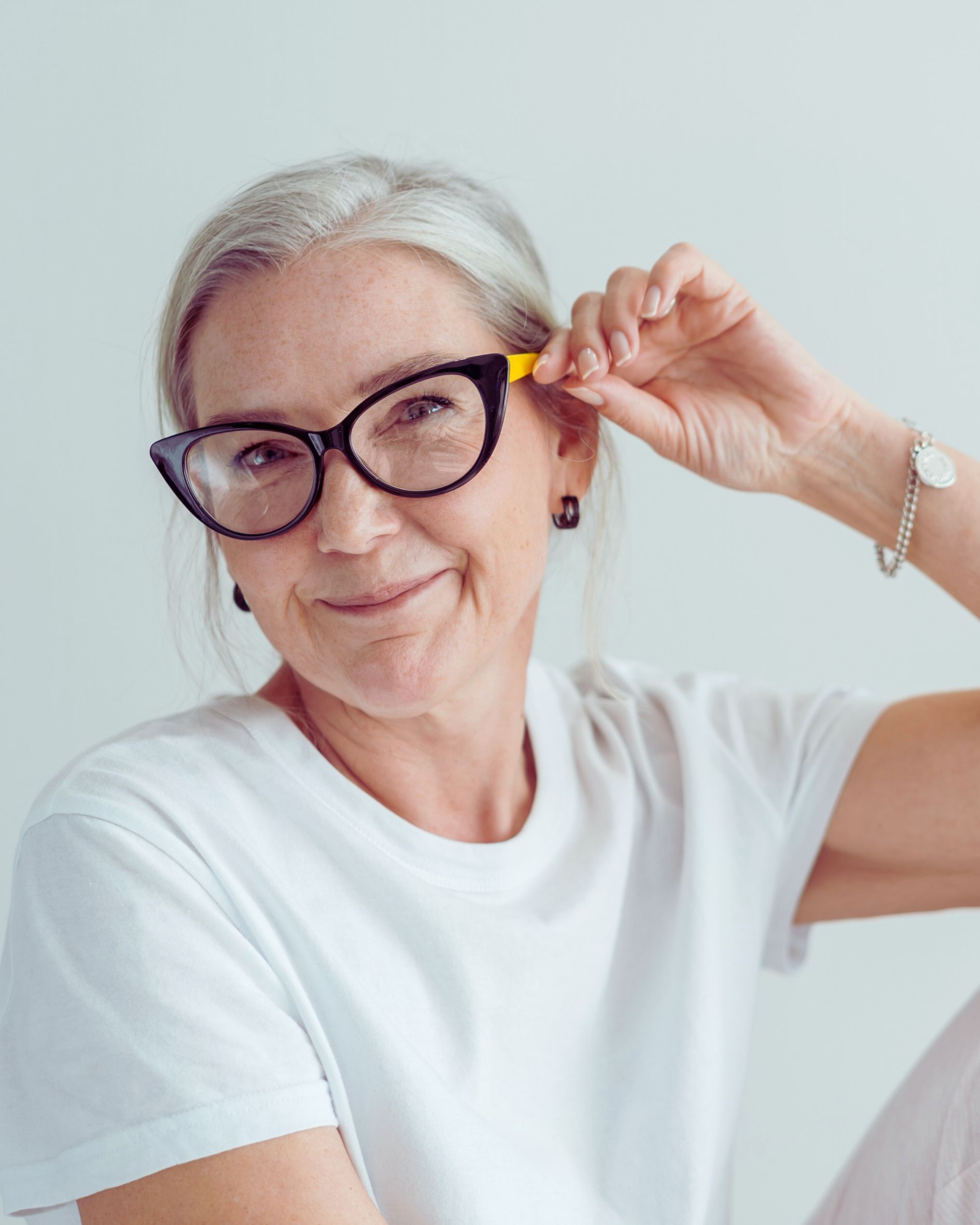 Woman with glasses, gray hair, holding yellow earplug, wearing white shirt.