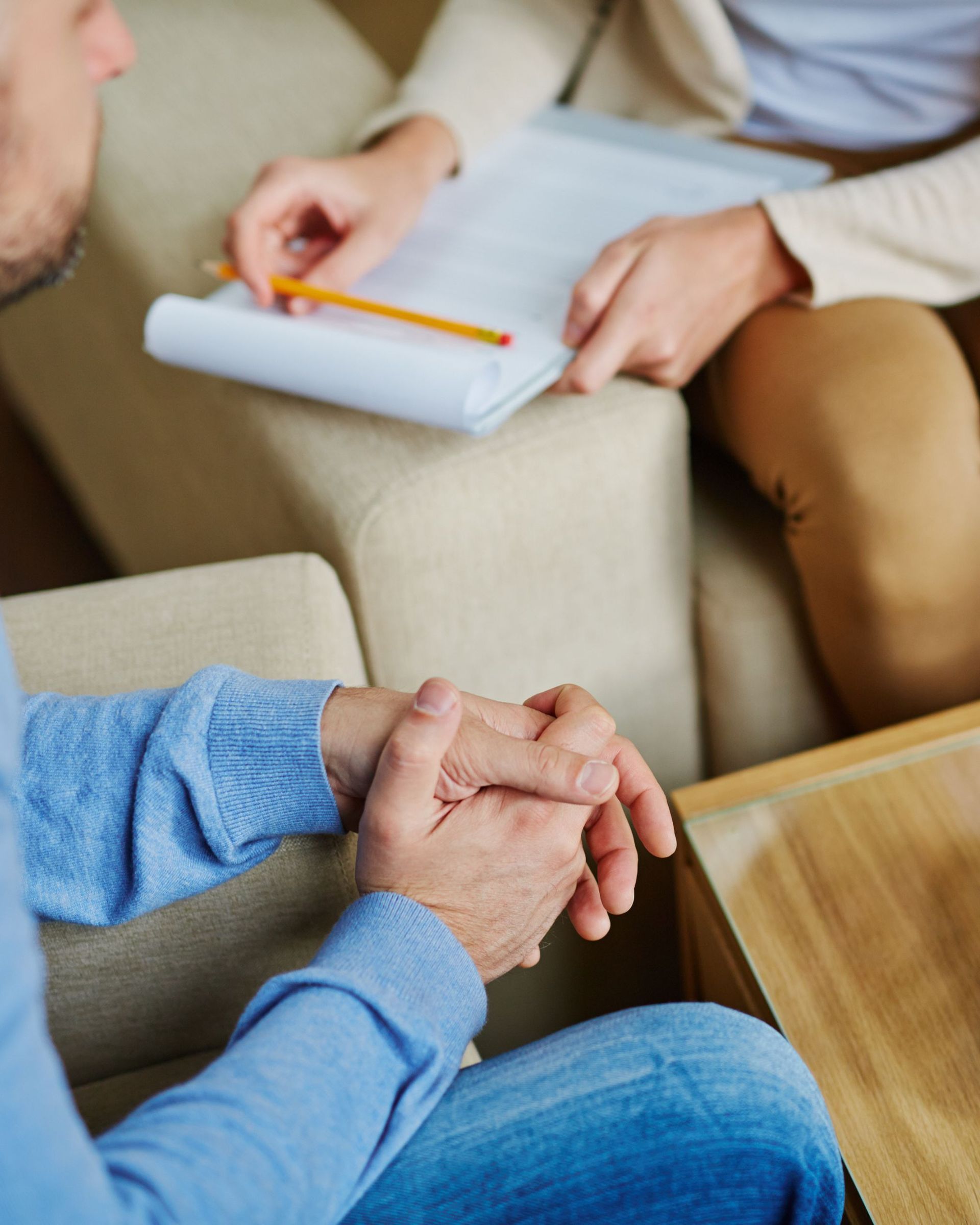 Man with folded hands in therapy session, counselor taking notes.