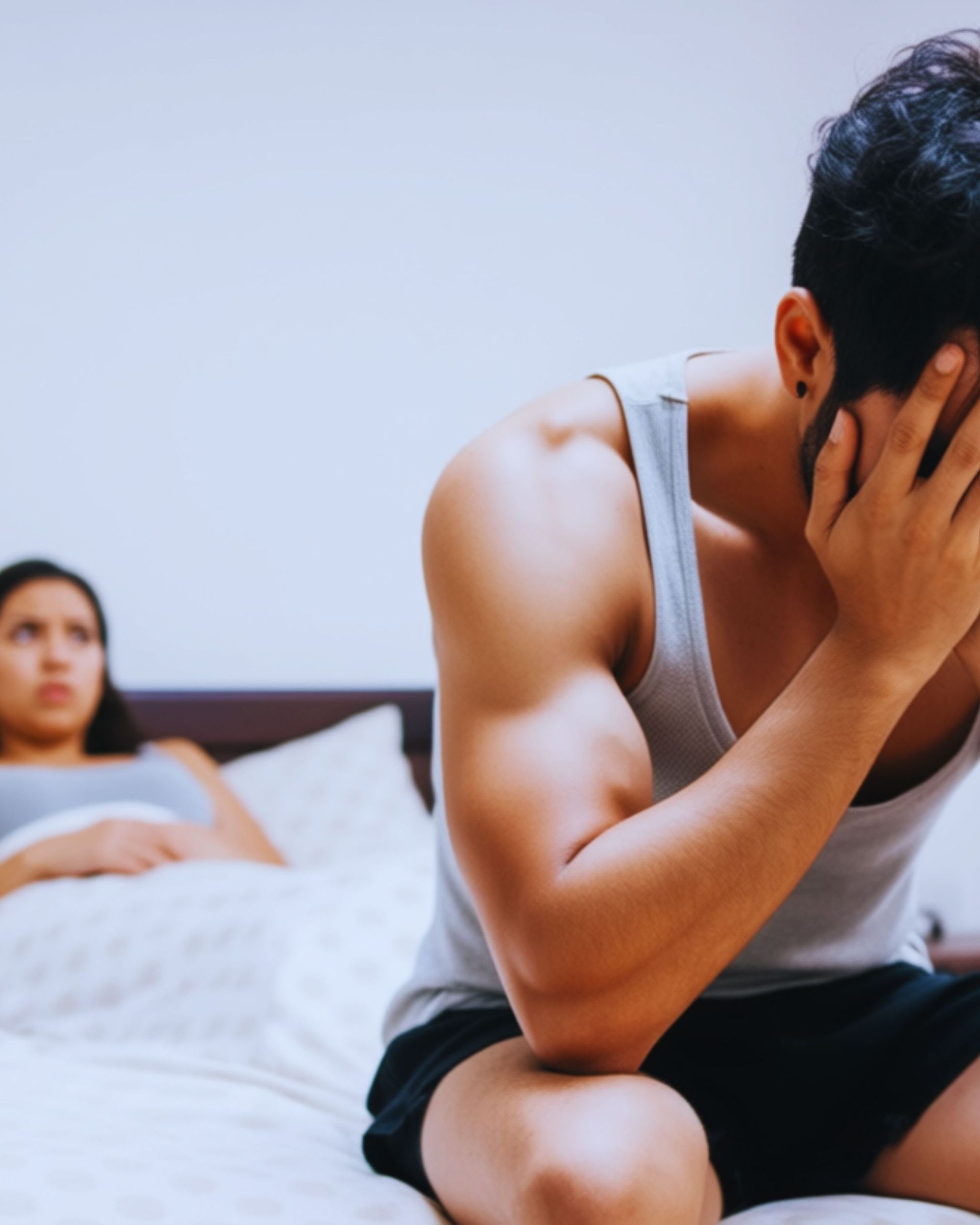 Man in a tank top with his face in his hands on a bed, woman in the background lying down.