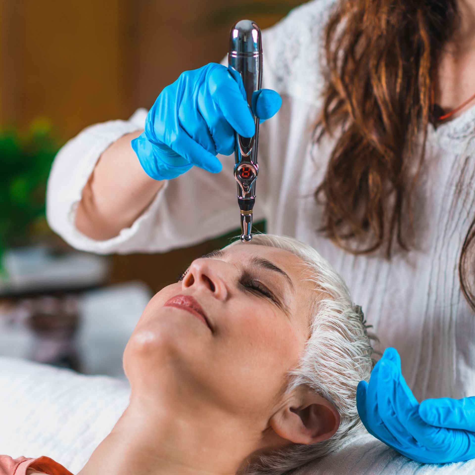 Person receiving head treatment with tool, gloved hands, eyes closed.