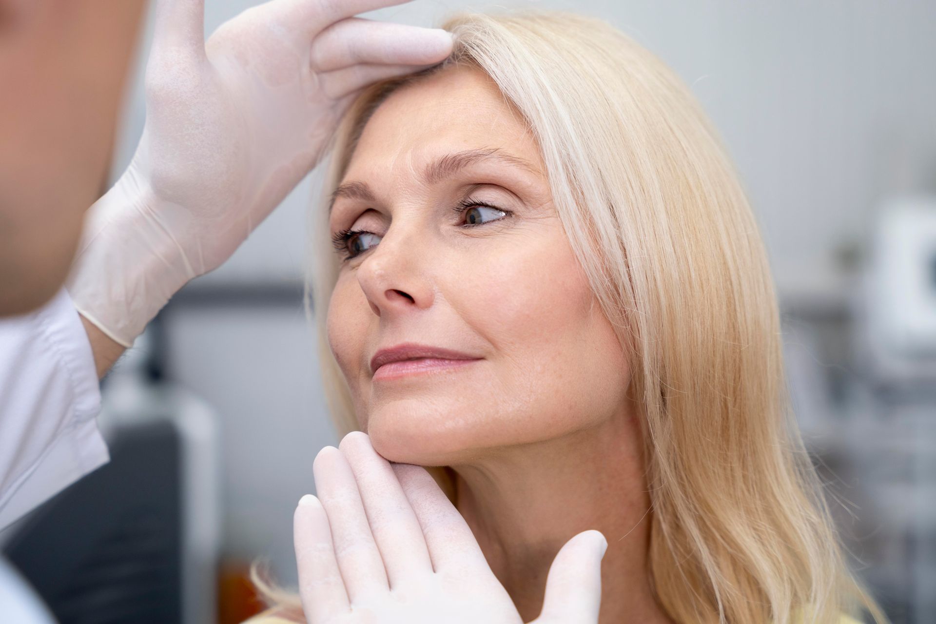 Doctor examining a woman's face, possibly for cosmetic procedure; hands are gloved, setting is a clinic.