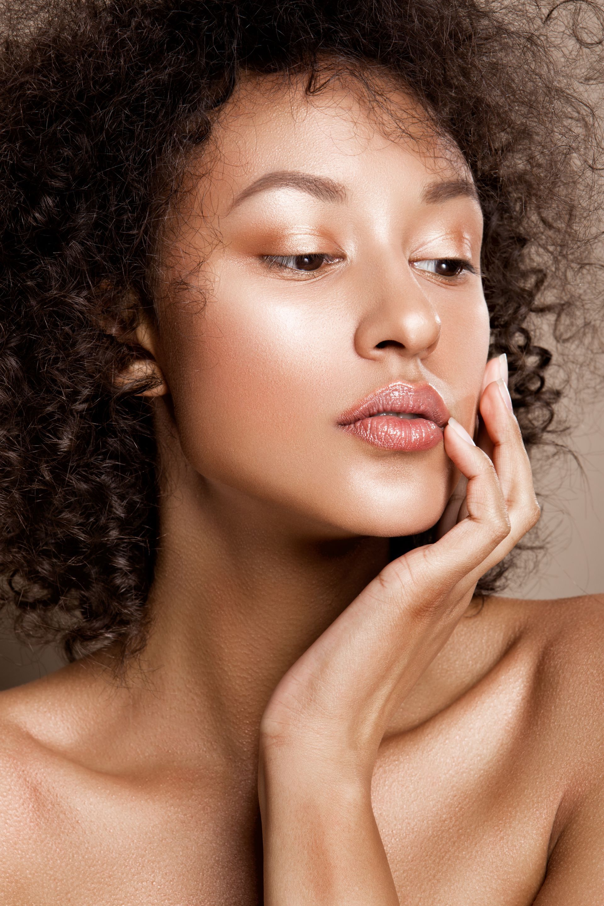 Woman with curly hair, hand to face, with highlighted skin, against a neutral background.