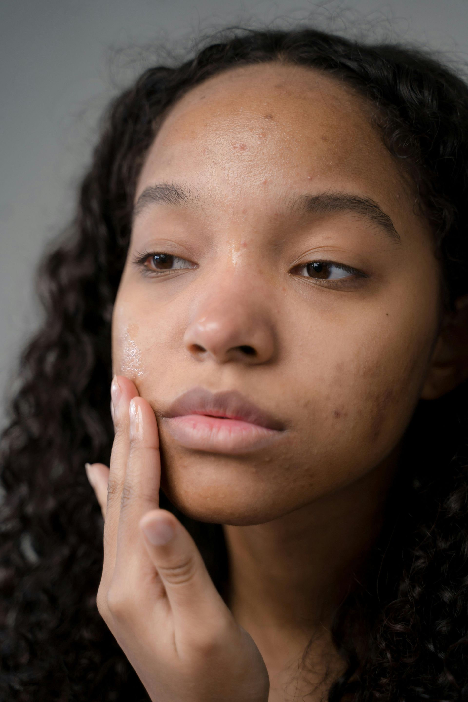 Woman applying skincare product to her face with hand, eyes closed, against a light gray background.