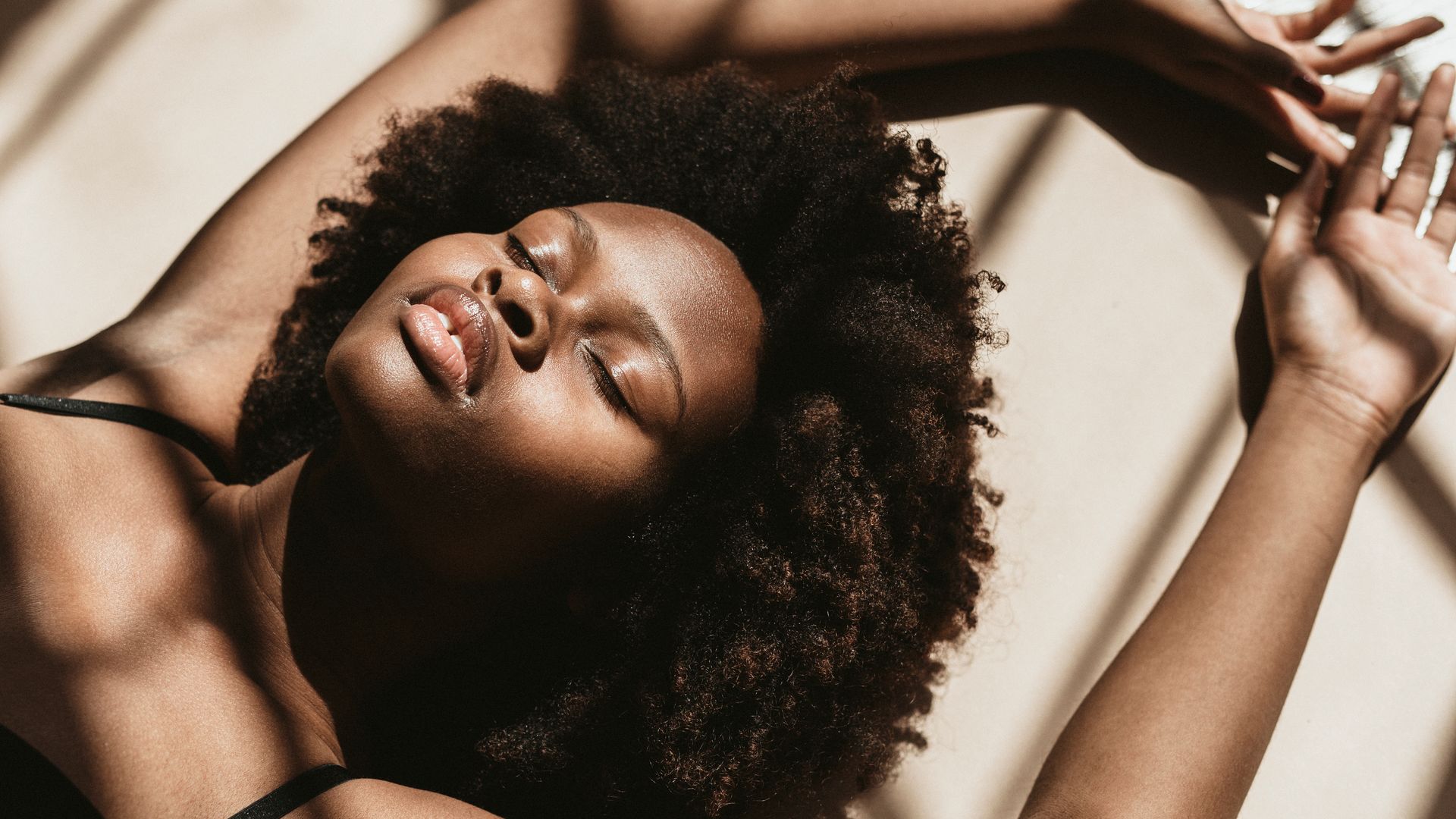 Woman with curly dark hair, eyes closed, arms raised, basking in sunlight.