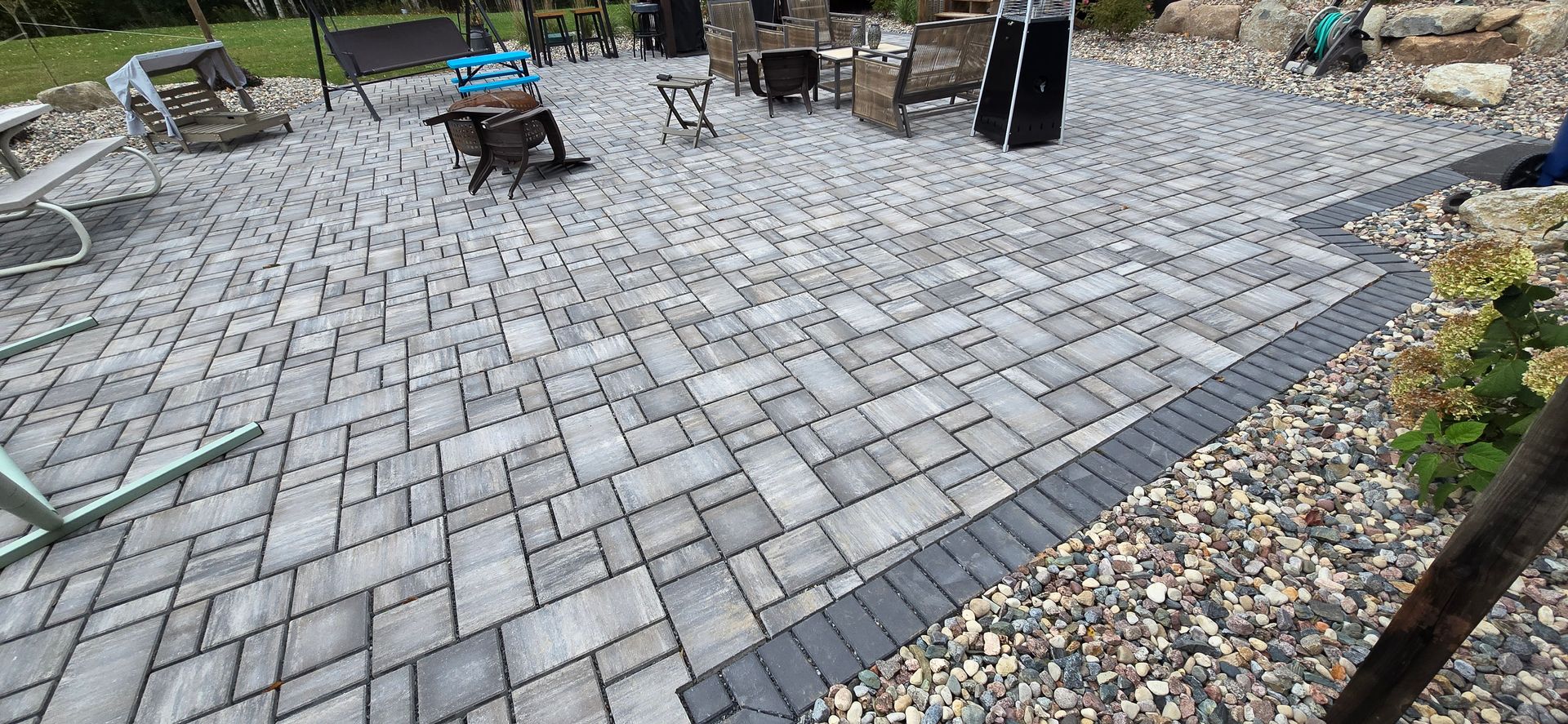 Gray stone patio with dark border next to gravel and a few chairs in a backyard setting.