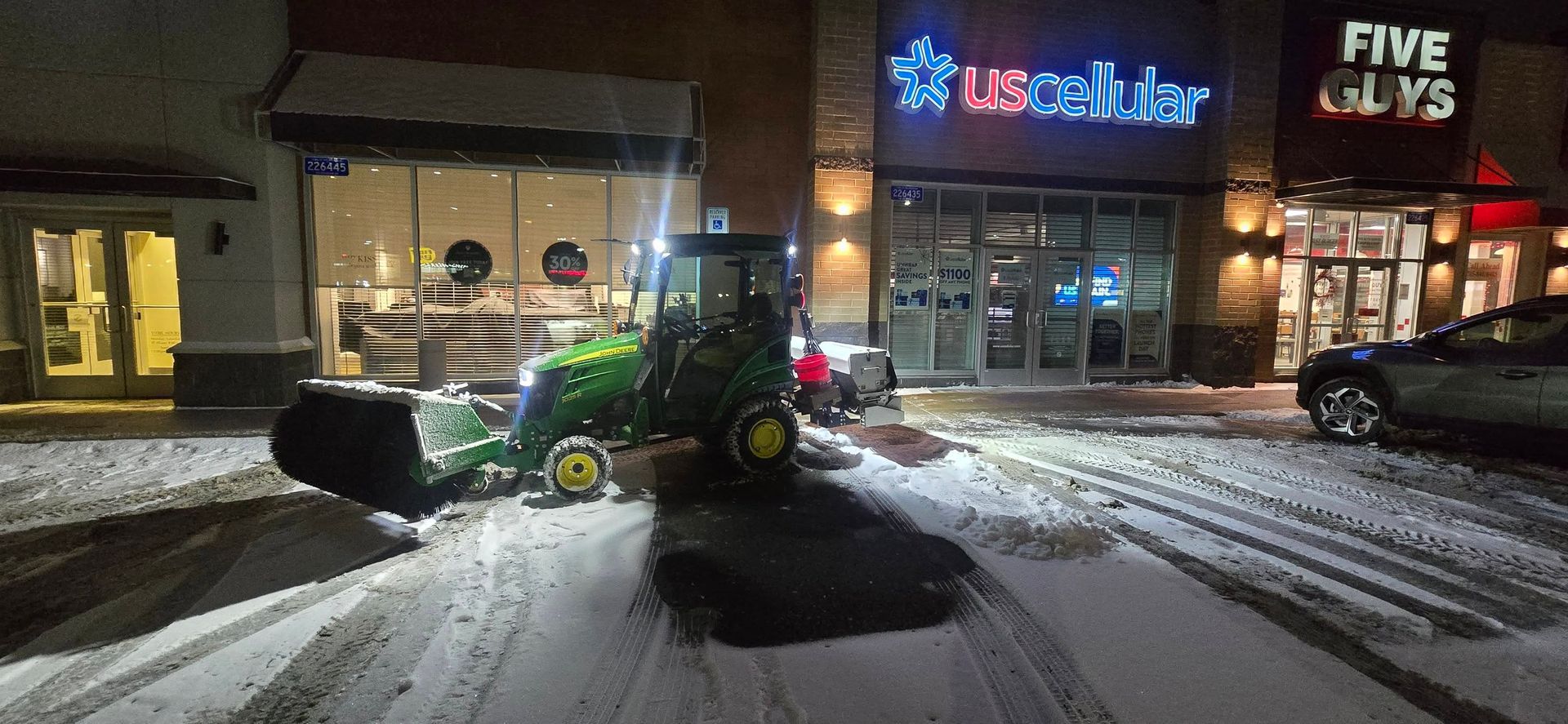 A green tractor plowing snow in front of a shopping center at night. Restaurants include US Cellular and Five Guys.