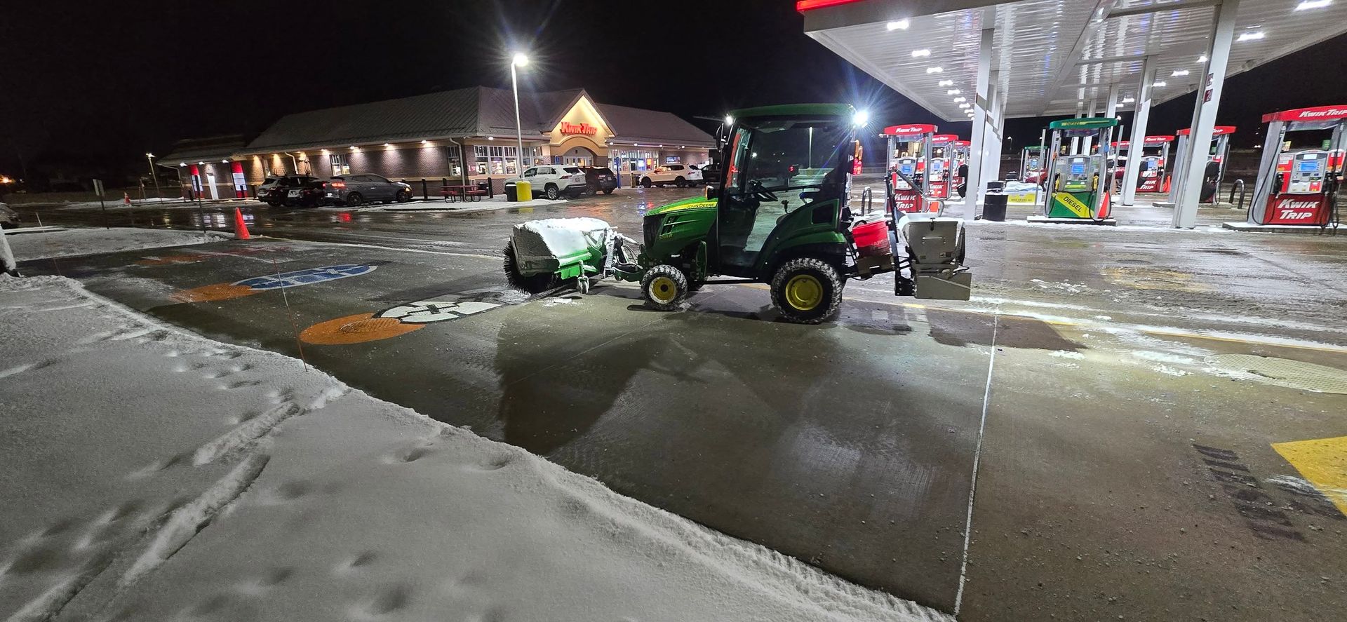A John Deere tractor clearing snow at a gas station at night.