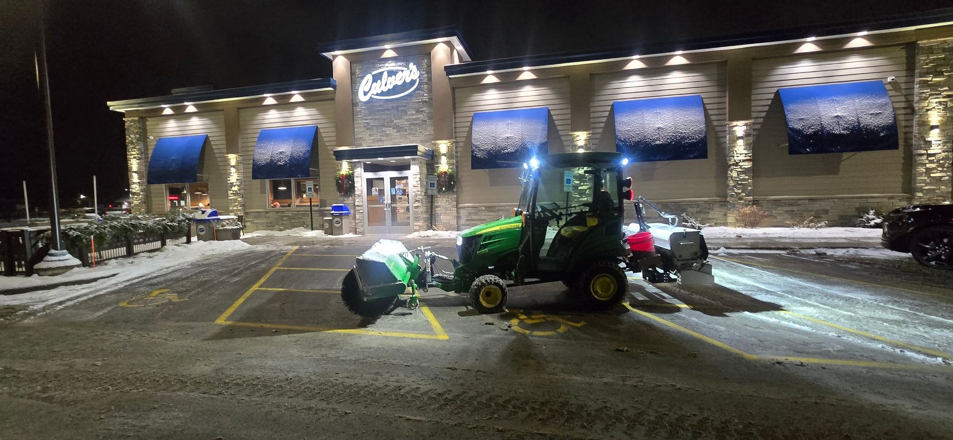 A snowplow clearing a parking lot in front of an exterior restaurant at night.
