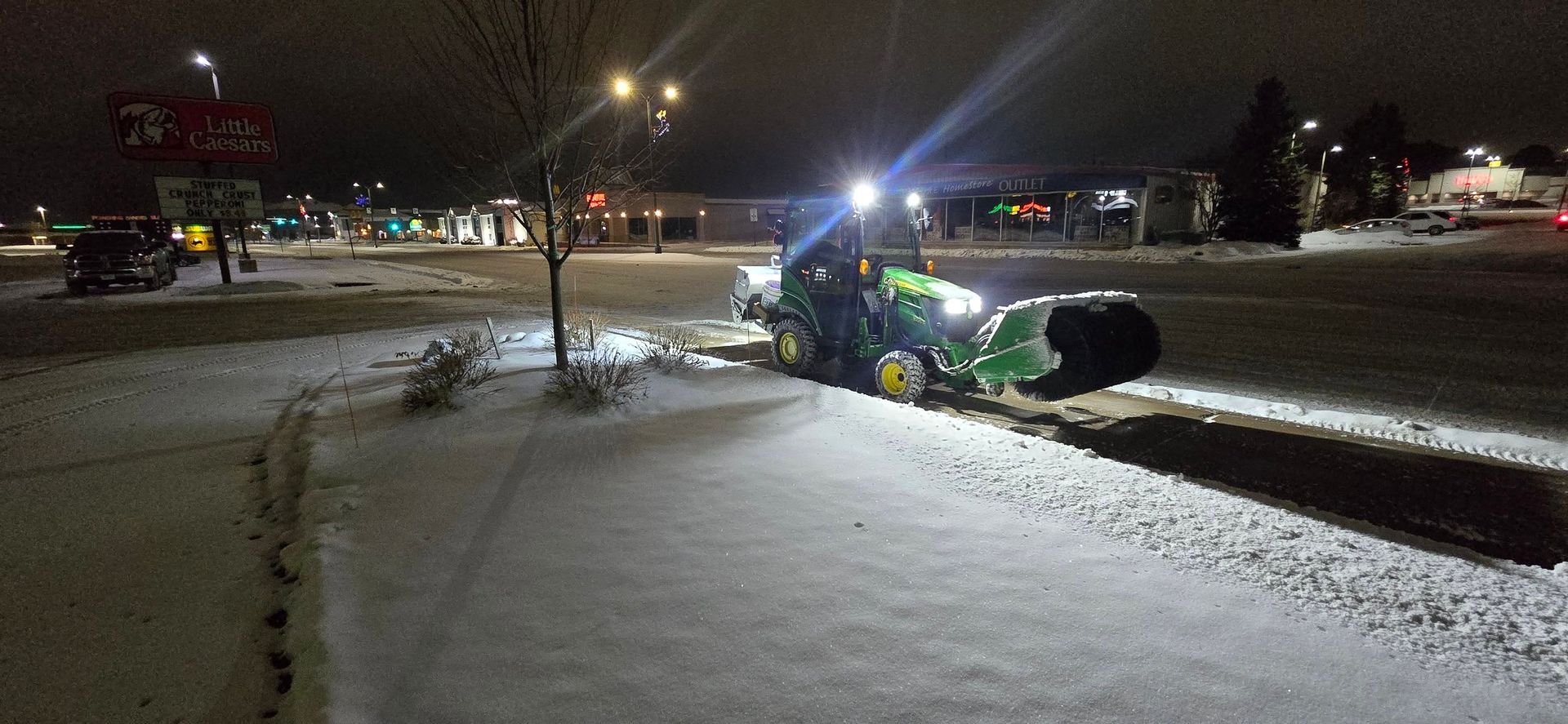 A John Deere tractor with snow blower clearing snow from a sidewalk at night.